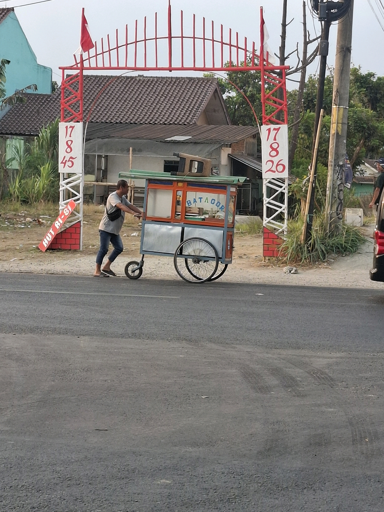 The image depicts an outdoor scene in the city of Karangtengah, Indonesia, featuring a man pushing a food cart under a decorative archway. The archway is red and white, with the numbers "17 8 45" on one side and "17 8 26" on the other, possibly commemorating Indonesian Independence Day. A red and white flag is attached to the top of the arch. The food cart is predominantly silver and green, with orange accents and signage that reads "BATAGOR". The man, wearing a gray t-shirt and blue jeans, is pushing the cart from the side, with his left hand on the handle. He is barefoot, wearing flip-flops. The background shows buildings with tiled roofs, lush green vegetation, and utility poles. The ground is paved with asphalt in the foreground, with a dirt and gravel shoulder on the right. Tire tracks are visible on the asphalt. The sky is overcast.
