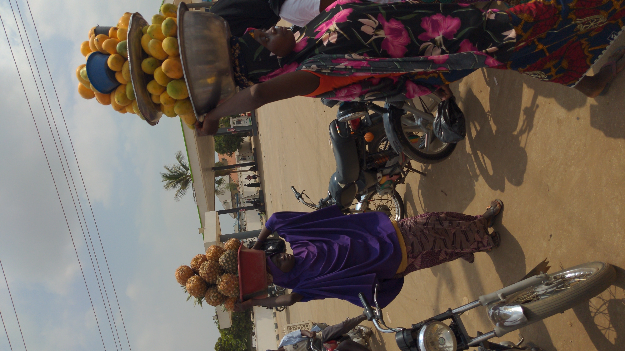 Two women are selling fruit on a street in Ado, Nigeria. One woman is carrying a stack of trays filled with mangoes on her head. The other woman is carrying a red tub filled with pineapples on her head. They are standing in a dusty area with motorcycles parked nearby, in front of a building with a green and white awning. Power lines run across the sky. The weather appears to be sunny. People can be seen in the background.