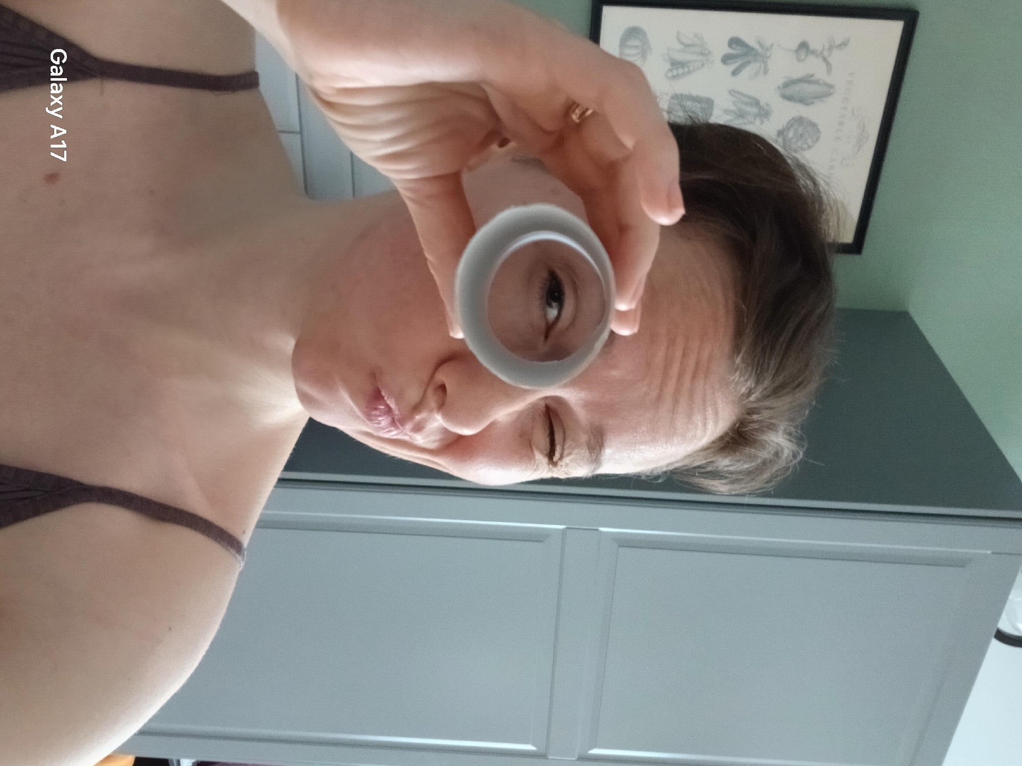 A woman is holding a cylindrical object up to her eye, looking through it. She is wearing a dark brown strap across her chest. Behind her are light grey cabinets and a framed picture of vegetables. The photo appears to have been taken indoors. There is no evidence to suggest this image was taken in Yekaterinburg, Russia.
