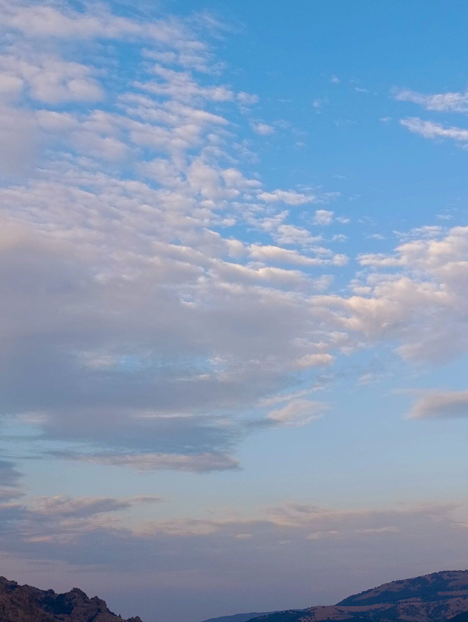The image depicts a vast expanse of a pale blue sky filled with scattered, wispy clouds. The clouds vary in density and color, with some appearing bright white and others tinged with soft pink and gray hues, suggesting either sunrise or sunset. In the lower portion of the image, a silhouetted mountain range with rugged peaks is visible, partly obscured by a soft haze. The terrain appears arid and sparsely vegetated. The overall scene is serene and natural, with no signs of human activity or artificial structures. This landscape is consistent with the natural environment surrounding Shiroko pole, Bulgaria, known for its hilly terrain and open skies.