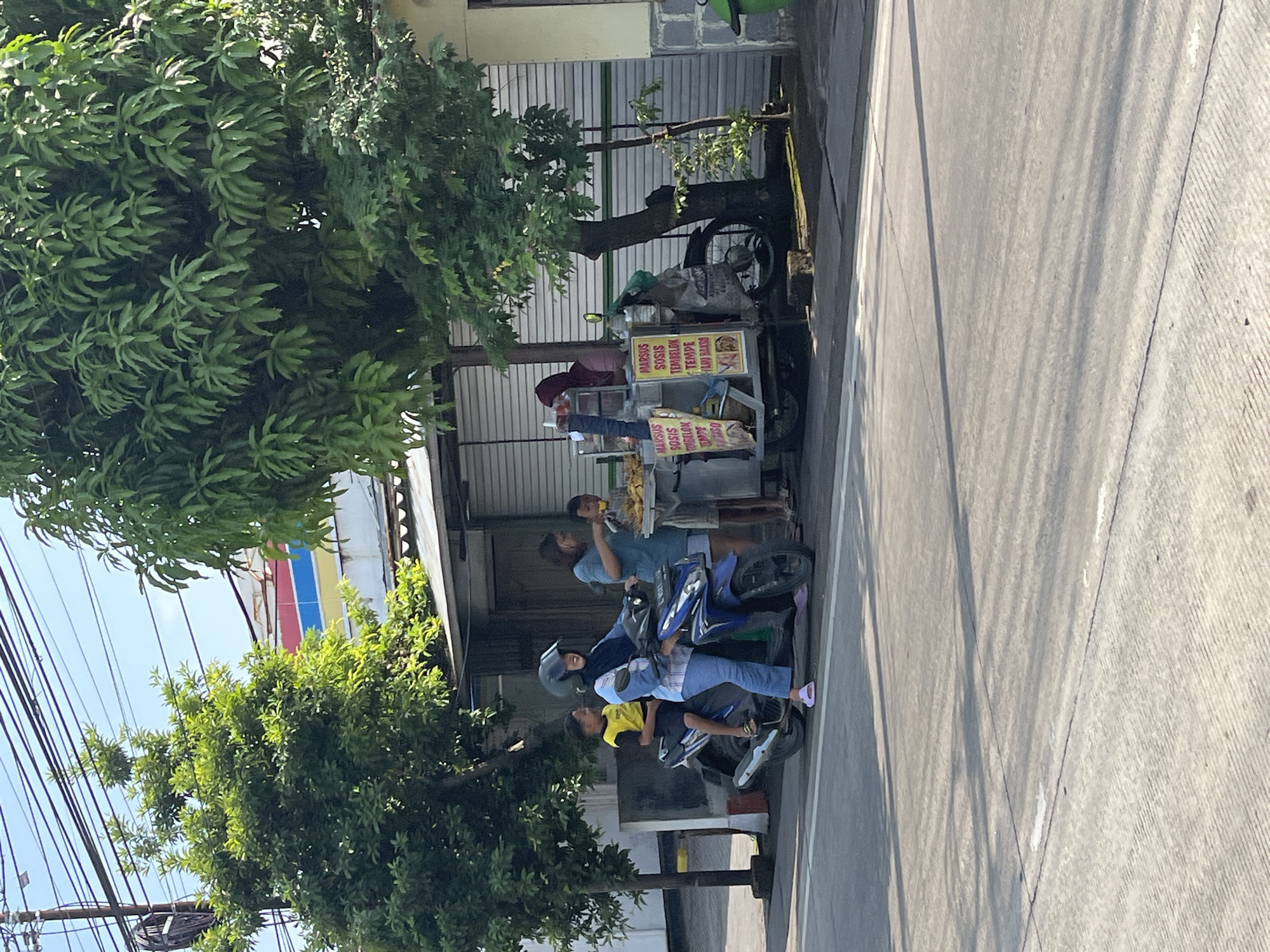 A woman in a blue hijab and a blue and white striped shirt, wearing blue jeans and pink flip-flops, is riding a blue scooter. A young boy in a yellow and black shirt is sitting behind her. They are on a paved road in front of a food stall. The food stall has a cart with several items displayed, including signs with Indonesian text such as "MARSUS SOSIS," "TEMBELOK," and "TEMPE." A woman in a light blue shirt and a girl in a red hijab are standing near the food stall. The background shows a building with a colorful facade, trees, and overhead wires. The scene appears to be in a street setting, likely in a commercial or residential area of Kajeksan, Indonesia, during daylight hours. The road surface exhibits shadows from trees and overhead structures.