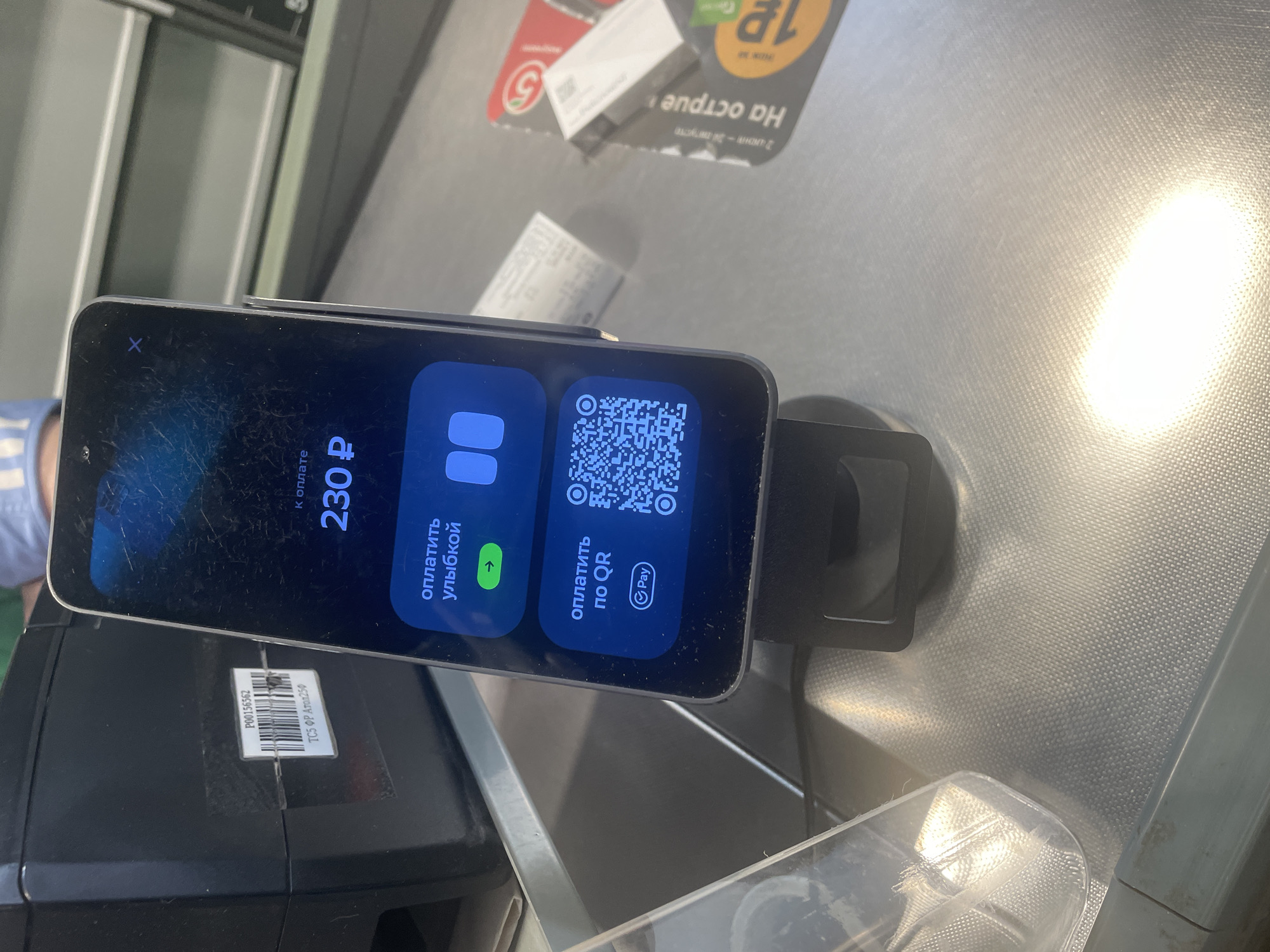 This media file captures a close-up, slightly angled view of a smartphone displaying a payment interface, likely at a checkout counter. The phone is propped up on a black stand, suggesting it's being used for a transaction. The screen shows a clear heading "к оплате" (to pay) followed by the amount "230 P" (230 rubles). Below this, two payment options are presented: "оплатить улыбкой" (pay with a smile) with an arrow pointing right, and "оплатить по QR" (pay by QR) with a QR code and a "Pay" icon. The phone's dark background is visible, with some scratches on its surface.
The background reveals a grey, textured surface, possibly a checkout counter at a retail store, with a bright light source reflecting off it, suggesting an indoor setting. Partially visible in the upper background are some retail displays and what appear to be price tags or promotional materials, one featuring the number "5" in a red circle, hinting at a supermarket or convenience store environment. Below the phone, a black electronic device, likely a cash register or payment terminal, is visible. A small sticker with a barcode and text "P00156562 ТСБ ФР Атол 250" is attached to this device. The overall scene suggests a modern payment process in progress, focusing on the technology used for contactless transactions. There are no people clearly visible, but the presence of a person's arm wearing a blue shirt is faintly discernible on the left side of the frame, indicating someone is operating the phone or the payment system. The lighting and reflection suggest it is daytime.