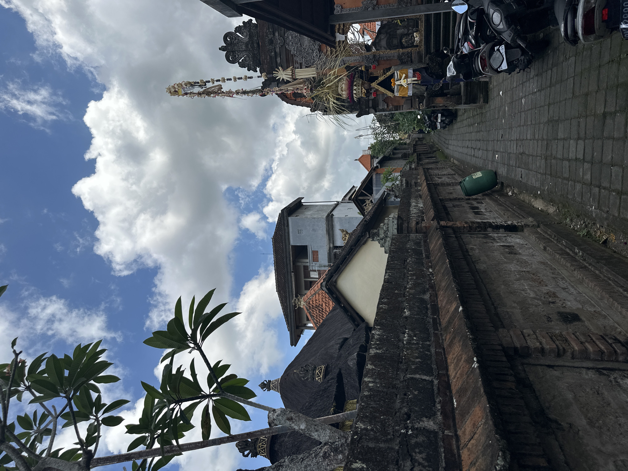 The image depicts a narrow, paved alleyway in the city of Ubud District, Indonesia. The alley is lined with traditional Balinese architecture, characterized by stone walls, thatched roofs, and ornate temple gateways adorned with festive decorations. Several motorcycles are parked along the right side of the alley. Lush green foliage from trees is visible in the upper left portion of the frame, and the sky is partly cloudy. The scene appears to be a quiet street within a residential or temple area.