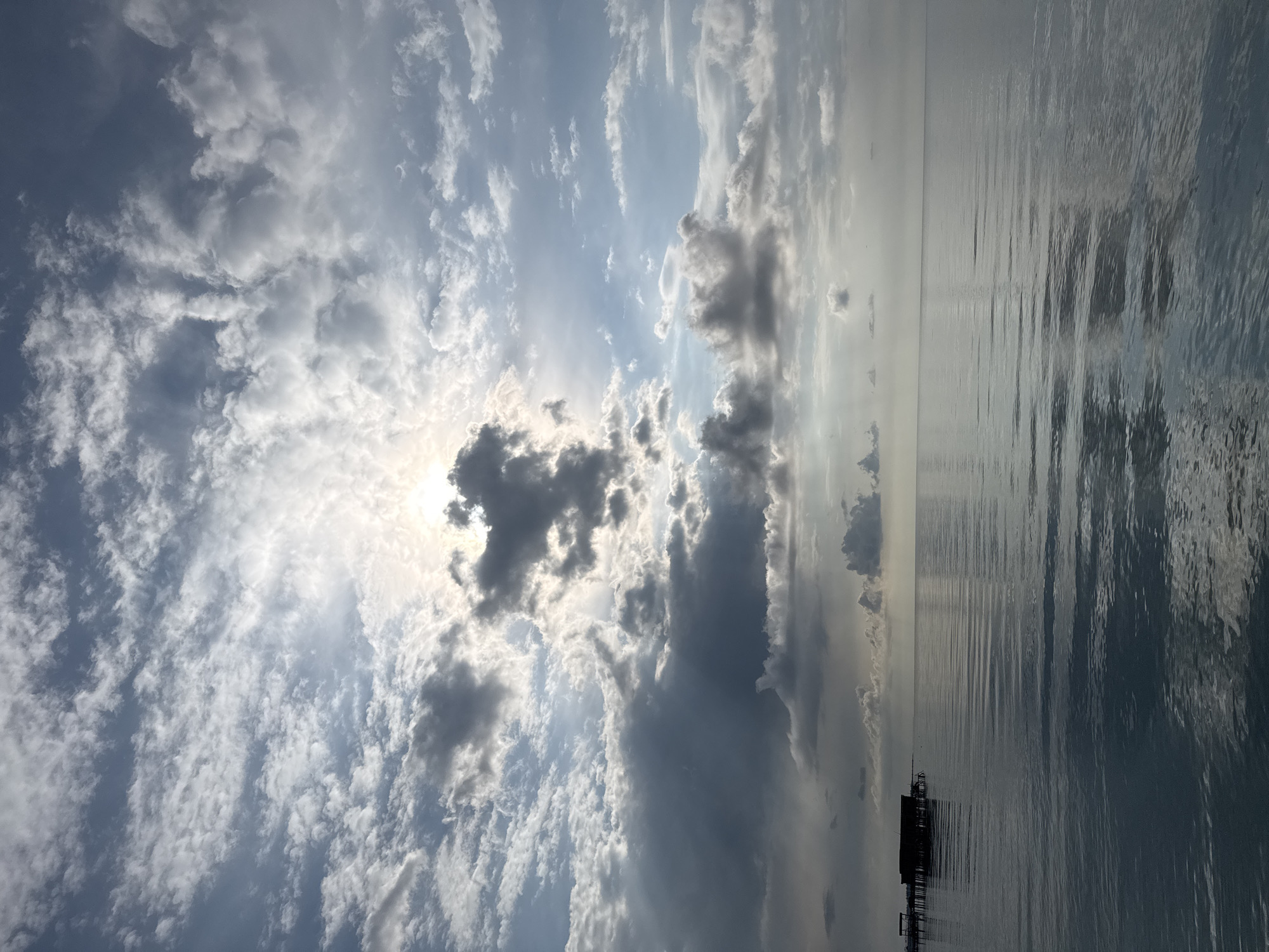 The image depicts a serene seascape with a partially cloudy sky and calm water. The sun is visible behind a cluster of dark clouds in the upper center, casting rays of light that illuminate the sky. The water's surface is smooth, with gentle ripples reflecting the sky's light and the silhouette of a small, dark structure on stilts in the distance on the left. The scene suggests a tranquil atmosphere. The location is stated as Sary-Oy, Kyrgyzstan.