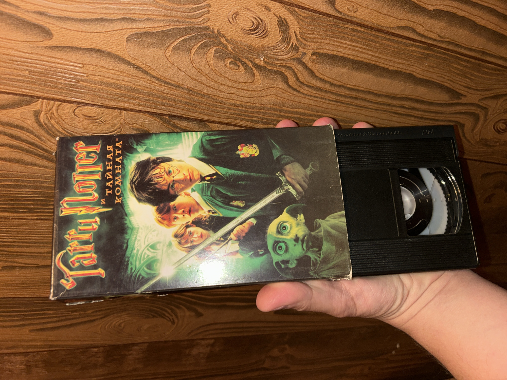 A hand is holding a VHS tape of "Harry Potter and the Chamber of Secrets" in Russian. The tape is positioned vertically against a wooden paneled wall. The VHS cover features characters from the movie, including Harry Potter, Ron Weasley, Hermione Granger, and Dobby. The background wall has a prominent wood grain pattern with vertical lines. The lighting is moderate, illuminating the tape and the hand holding it. There are no visible signs of the city of Lyubertsy, Russia, in the image.