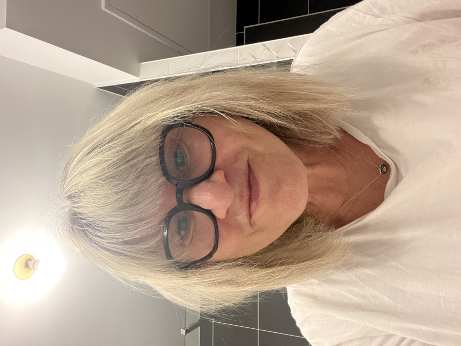 A woman with shoulder-length blonde hair and bangs is wearing large, black-rimmed glasses. She wears a simple white t-shirt and a delicate necklace. Her skin shows signs of aging. The photo appears to be taken in a bathroom setting with white walls, dark tile and a light fixture visible in the background. The photo is slightly tilted, giving the impression that it was taken at an odd angle.