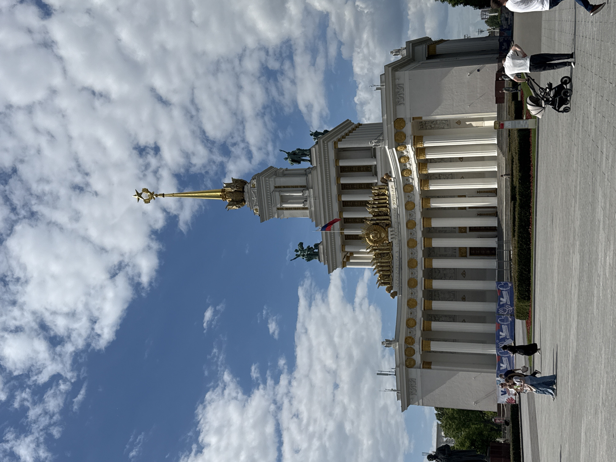 A grand, ornate building with classical architectural elements stands prominently under a bright blue sky dotted with scattered white clouds. The structure features white walls, numerous tall white columns, and extensive golden decorative elements, including a golden frieze with visible Cyrillic text, which appears to include "СССР" (USSR). A tall, golden spire, topped with a star, rises from the center of the building. Statues depicting human figures are positioned on the roofline. A Russian flag flies from a pole attached to the building.
In the foreground, a wide, paved plaza stretches out, with landscaped green hedges flanking the path. Several people are visible on the plaza; one individual is pushing a baby stroller, accompanied by another person, while a few other figures walk further in the distance. The scene is set during daytime under sunny weather conditions, suggesting a public park or exhibition ground. The architectural style, including the star-topped spire and Soviet-era inscription, strongly indicates the location as the VDNKh (Exhibition of Achievements of National Economy) in Moscow, Russia. A blue and white banner with indistinct text and graphics is visible near the base of the building.
