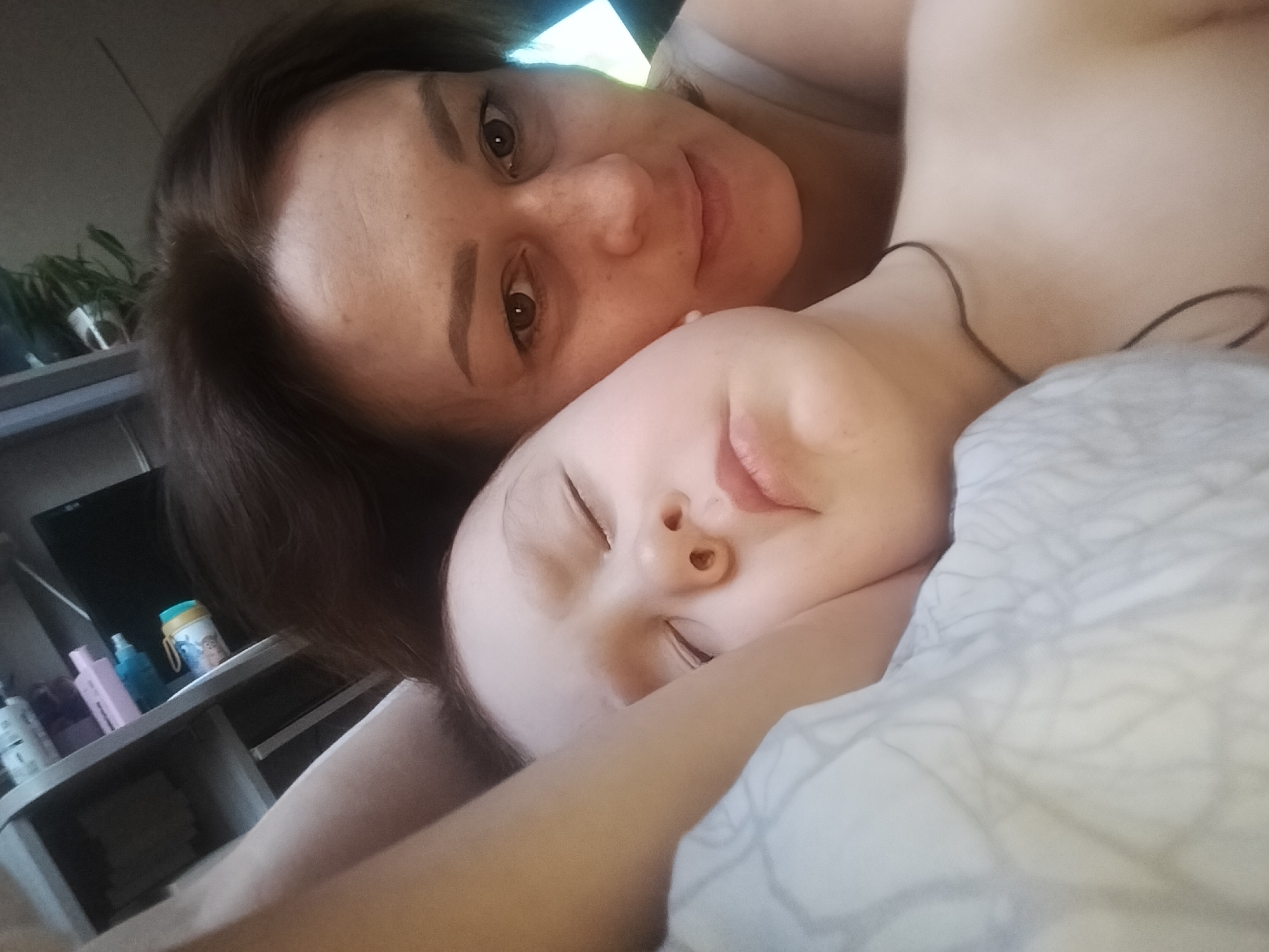 An intimate, close-up photograph captures an adult woman and a young child resting together, likely in a bed, in Chaikovsky, Russia. The image is taken from a slightly upward angle, giving the impression of a selfie or a shot taken by someone lying down with the subjects.
The adult woman, positioned in the upper part of the frame, has dark brown hair and brown eyes. She looks directly at the camera with a gentle, soft smile. Her skin is fair, with a scattering of freckles visible across her nose and cheeks. Her head is tilted slightly, and her shoulder is partially visible.
Nestled beside the woman, in the lower and central part of the frame, is a child with fair skin. The child's eyes are closed or nearly closed, suggesting they are sleeping or deeply relaxed. Their small nose and delicate lips are visible, conveying a peaceful demeanor. The child's arm is partially visible, resting on what appears to be white bedding with a subtle grey pattern. A thin dark string or necklace is visible on the child's neck.
In the background, a shelf or dresser can be seen, holding various items. These include several bottles, one of which appears to be a light pink dispenser, and another a blue one. A distinctive light blue cup with a cartoon character, possibly Stitch from Lilo & Stitch, is also visible. A dark screen, potentially a television, is behind these items. In the upper left corner, a green leafy houseplant is visible, adding a touch of natural element to the indoor setting. The lighting is soft and ambient, contributing to the calm and tender atmosphere of the scene. The overall impression is one of warmth, comfort, and maternal affection.