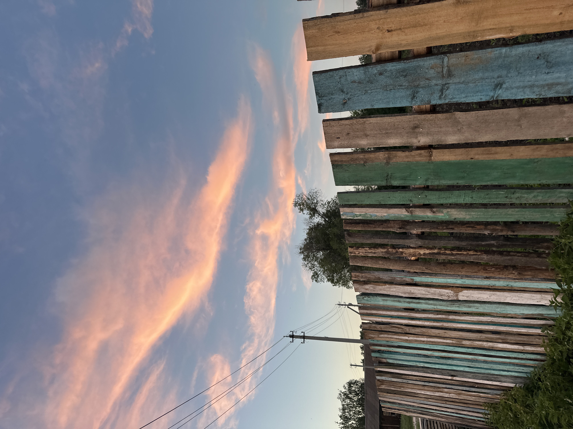 A low-angle outdoor shot shows a rustic, weathered wooden fence against a soft pastel sky at dusk. The sky is predominantly blue with streaks of pink and orange clouds, suggesting the setting sun. Three dark power lines stretch diagonally across the upper left quadrant of the frame, connected to a utility pole visible in the middle ground. Beyond the fence, trees and a dark-colored structure are dimly visible. The fence itself is constructed from vertical wooden planks of varying widths and conditions. Some planks are painted in faded colors of teal, green, and white, while others appear to be unpainted wood in shades of brown and gray. The foreground is occupied by lush green foliage. The scene evokes a rural or suburban setting during twilight.