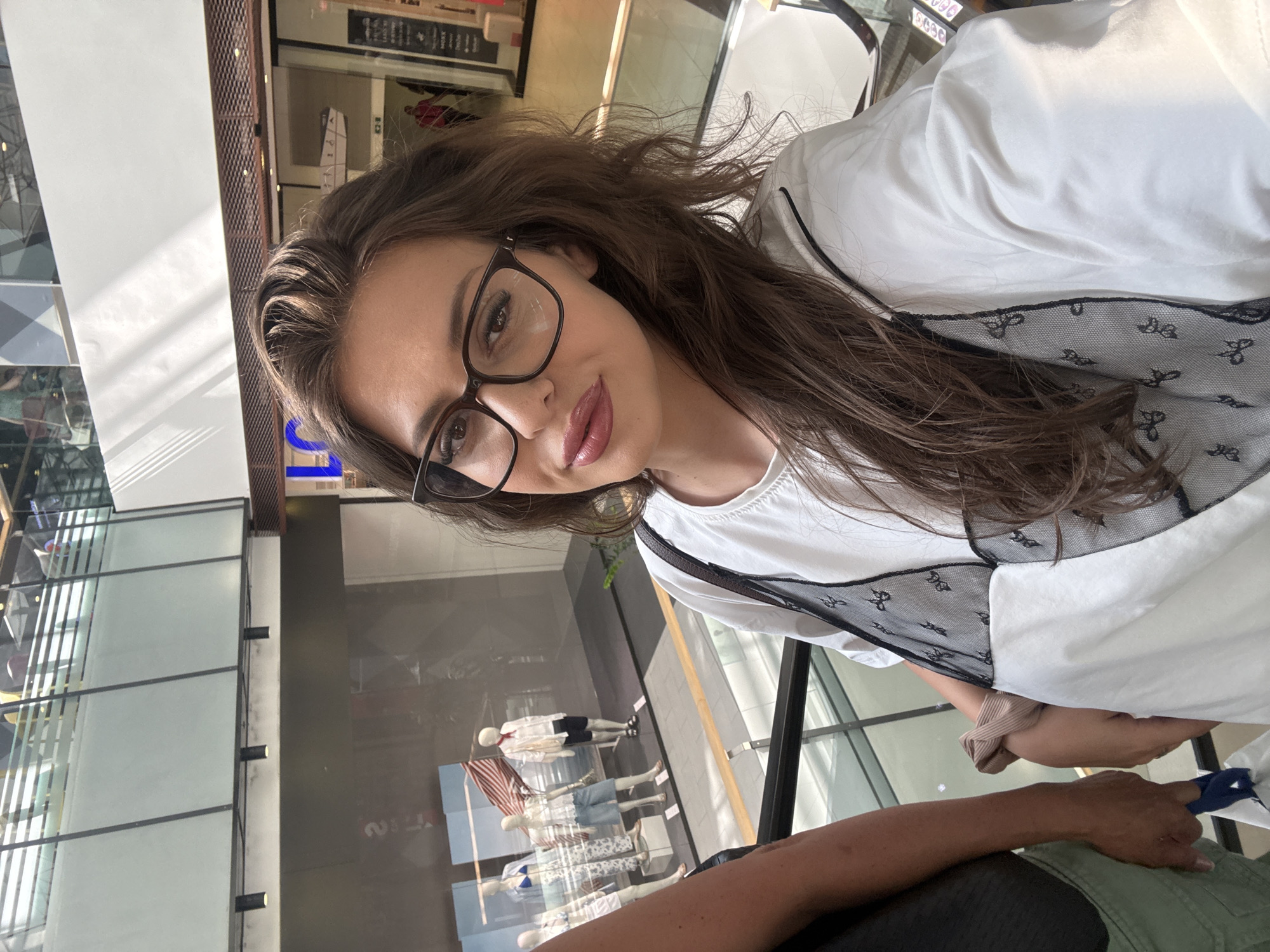 A young woman with long, brown hair and glasses is taking a selfie on an escalator in a modern shopping mall. She is wearing a white t-shirt with a black lace detail. To her left, a person's arm and torso are visible, with their hand holding a blue shopping bag. In the background, mannequins in a store window display summer clothing. The mall features multiple levels with glass railings and natural light streaming in from above. The woman appears to be in the center of the image, looking directly at the camera with a slight smile. The overall impression is a casual outing in an urban shopping environment.