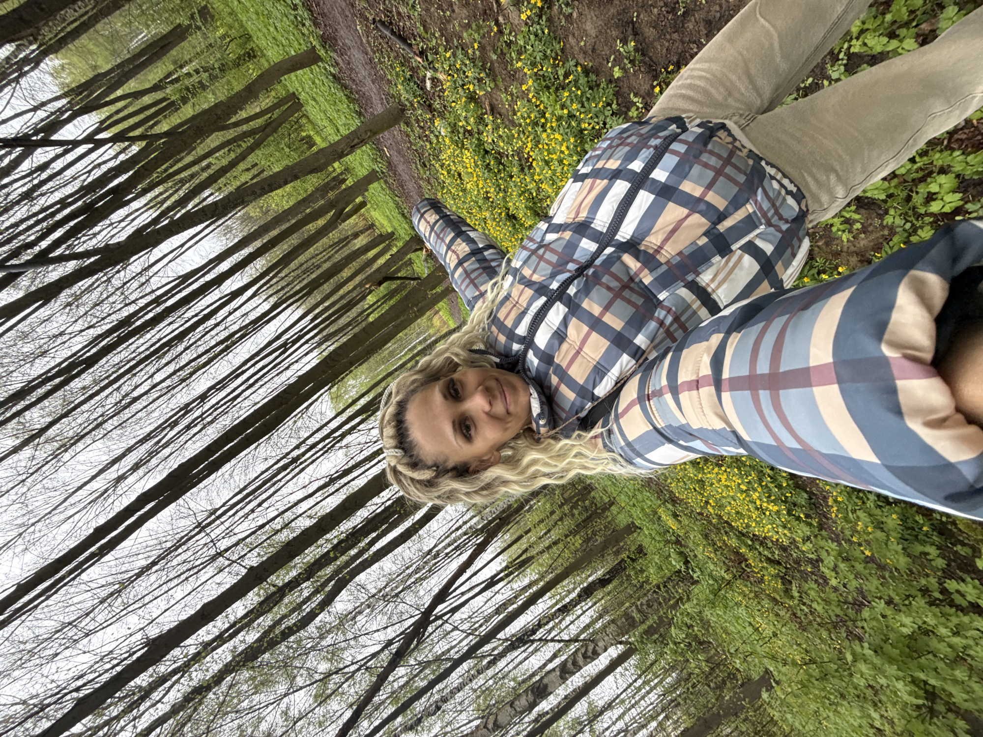 The image shows a woman in a plaid jacket and khaki pants lying on the ground in a forest or wooded area. The woman is fair-skinned with blonde, wavy hair. The ground is covered in green foliage and some small yellow flowers. Tall, bare trees dominate the background, creating a vertical perspective. A narrow dirt path is visible behind the woman. The location is likely a forested area. Given the location of Yaroslavl, Russia, it is likely in the surrounding woodlands or forest.
