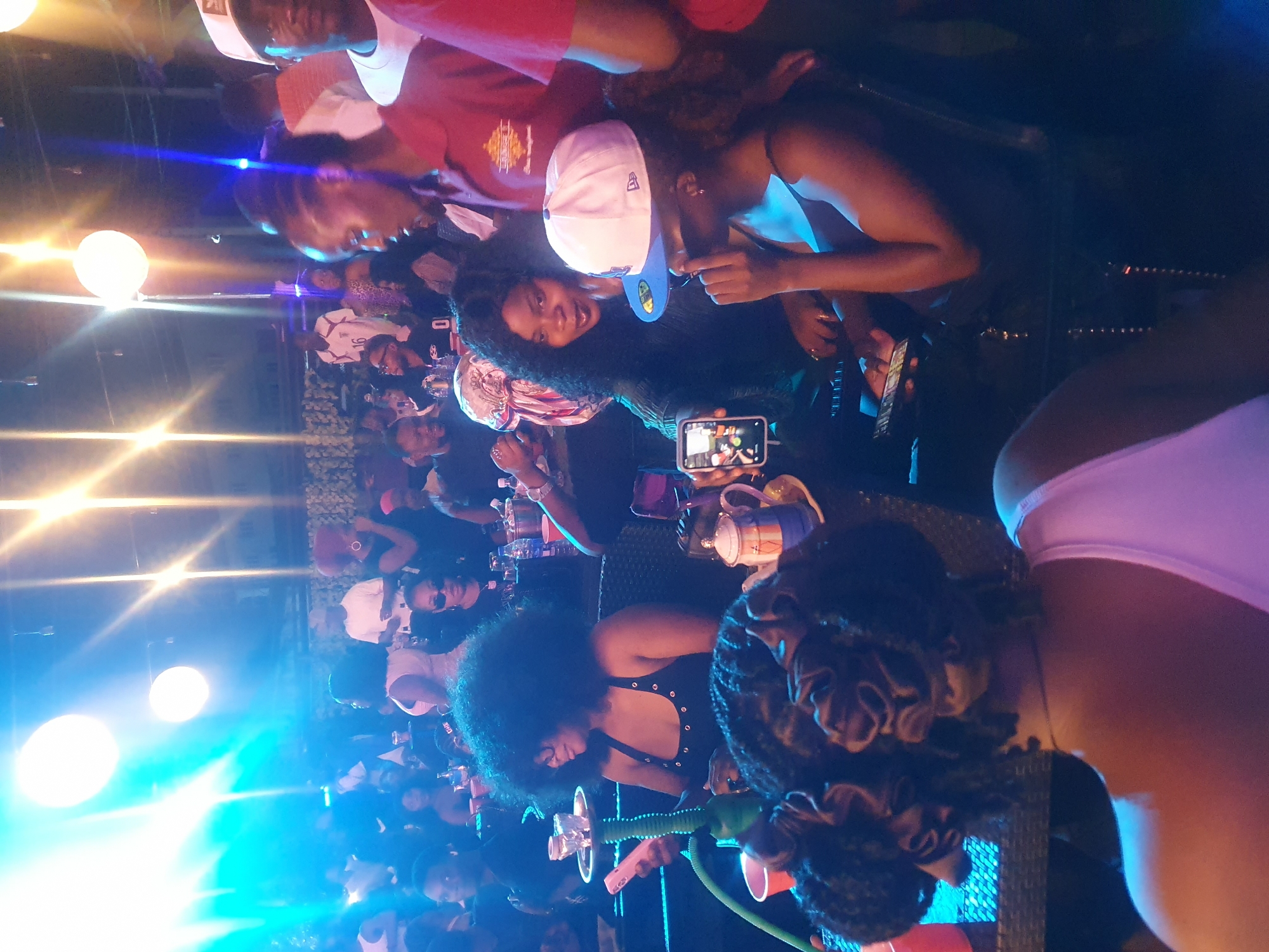 A nighttime scene in Lokoja, Nigeria depicts a crowded gathering. Individuals of varied skin tones are present, some wearing casual attire like T-shirts and baseball caps. One person is holding a smartphone, seemingly recording or taking pictures. People are at tables with cups and bottles, and at least one person is holding a hookah. The lighting is bright, with spherical light fixtures visible, and bluish tones dominate the scene.