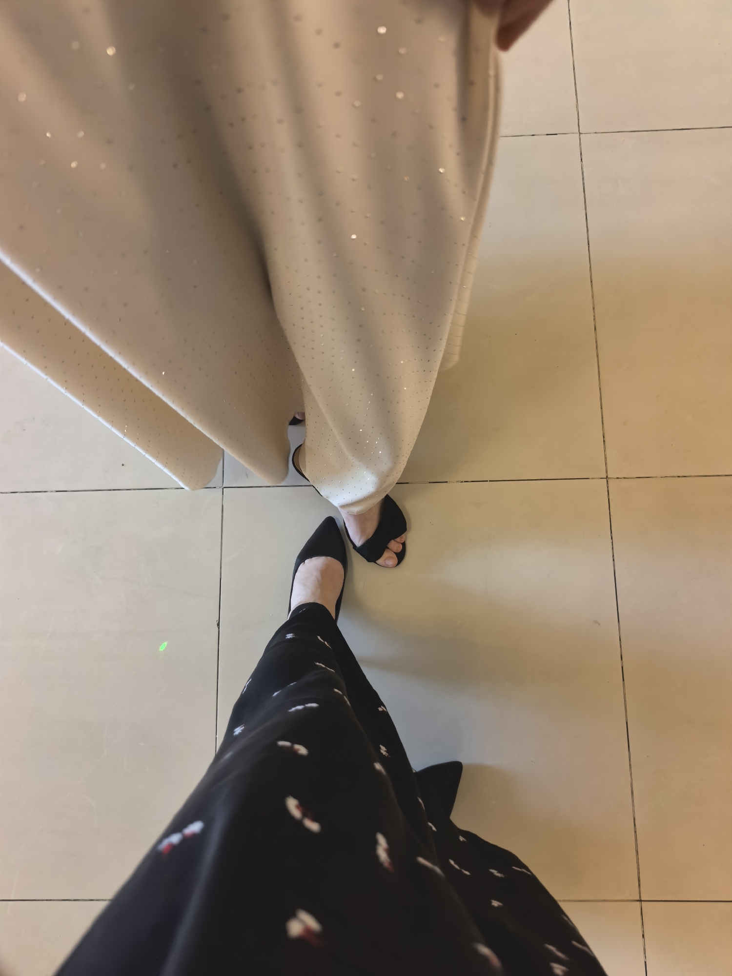Two individuals are standing on a tiled floor. The person in the foreground is wearing a black dress with a white floral pattern and black pointed-toe heels. The second person, positioned further back and to the right, is wearing a light-colored, floor-length dress adorned with small, shimmering dots and black open-toed heels. The floor is composed of large, square tiles with grout lines. The scene is lit from above, casting subtle shadows.