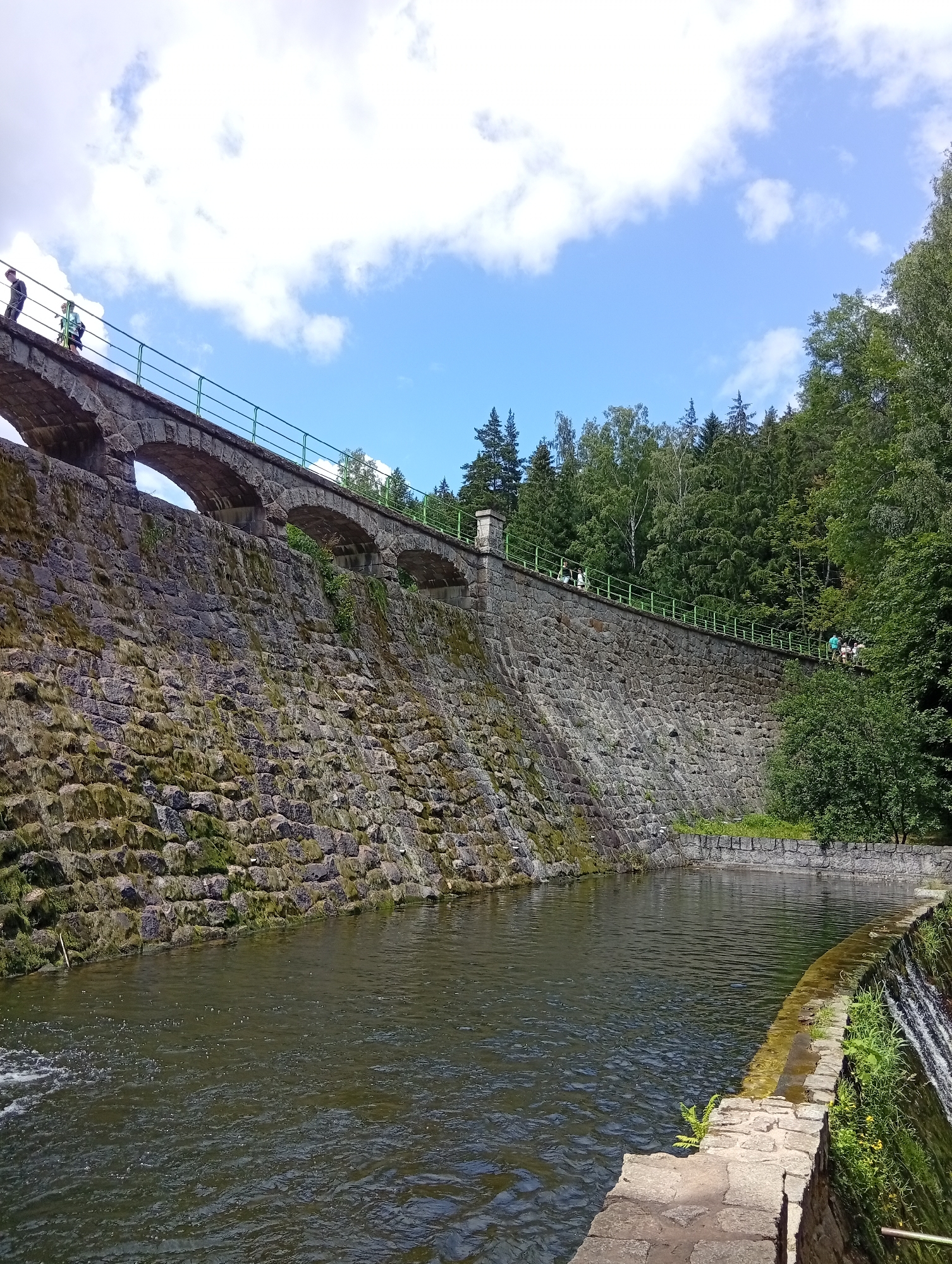 This is an image of the Karpacz Dam in Karpacz, Poland. The dam is made of large, rough-hewn stones and is covered in patches of moss. Above the dam, there is a walkway with a green railing, and several people are walking along it. The walkway is supported by arched stone structures that are part of the dam's construction. The dam holds back a body of dark, rippling water that flows away from the viewer on the right side of the image, where there is a small waterfall. Lush green trees and a bright blue sky with white clouds are visible behind the dam.