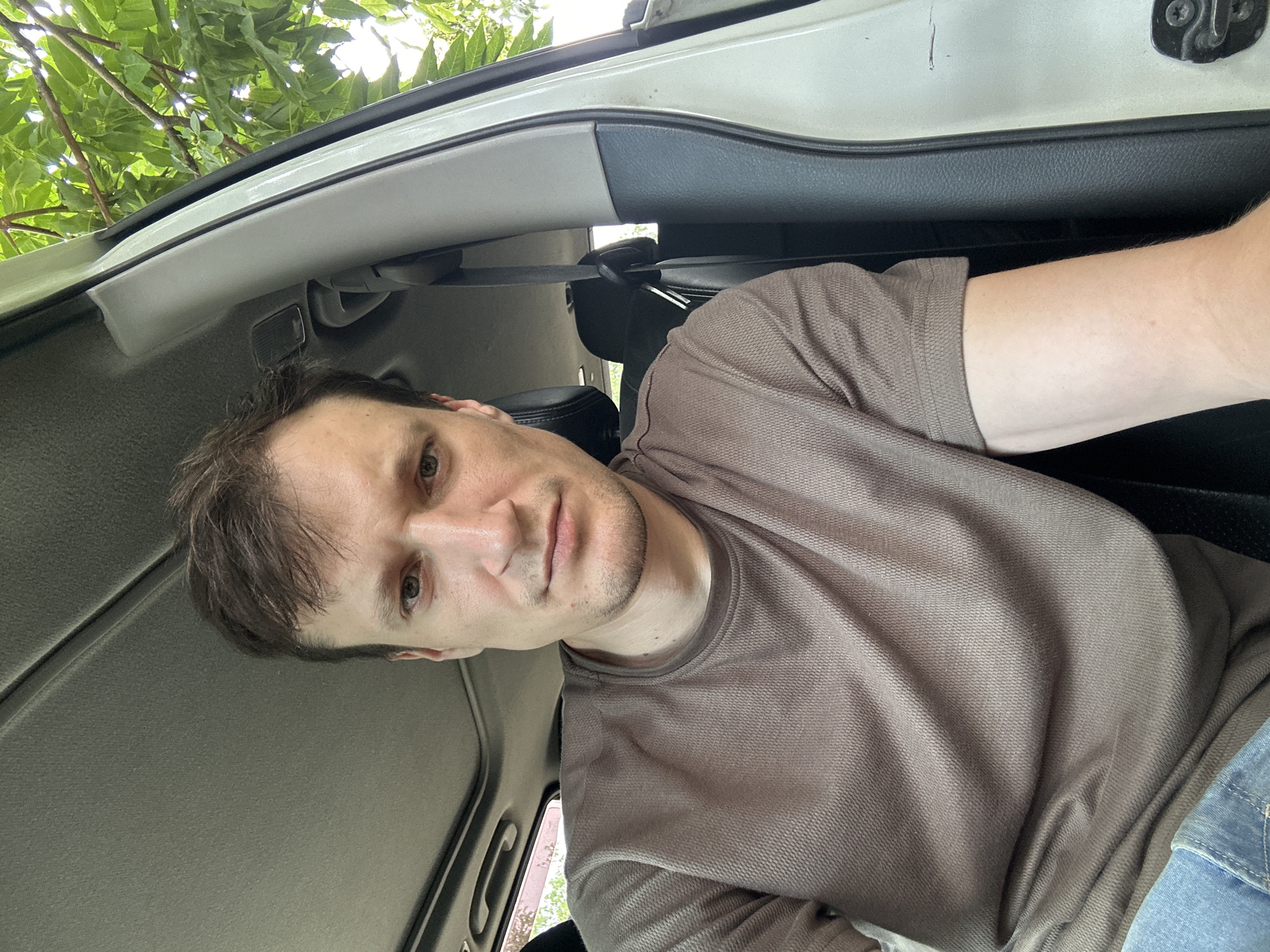 A medium shot shows a man sitting in the driver's seat of a vehicle, looking towards the camera. He has short, brown hair and is wearing a brown t-shirt and blue jeans. The interior of the car is visible, including the car's roof with a sunroof, the dashboard, and the driver's seat. Sunlight filters through the trees outside, casting shadows on the car's interior. The man's facial expression is neutral. The image appears to have been taken in Makiv, Ukraine.