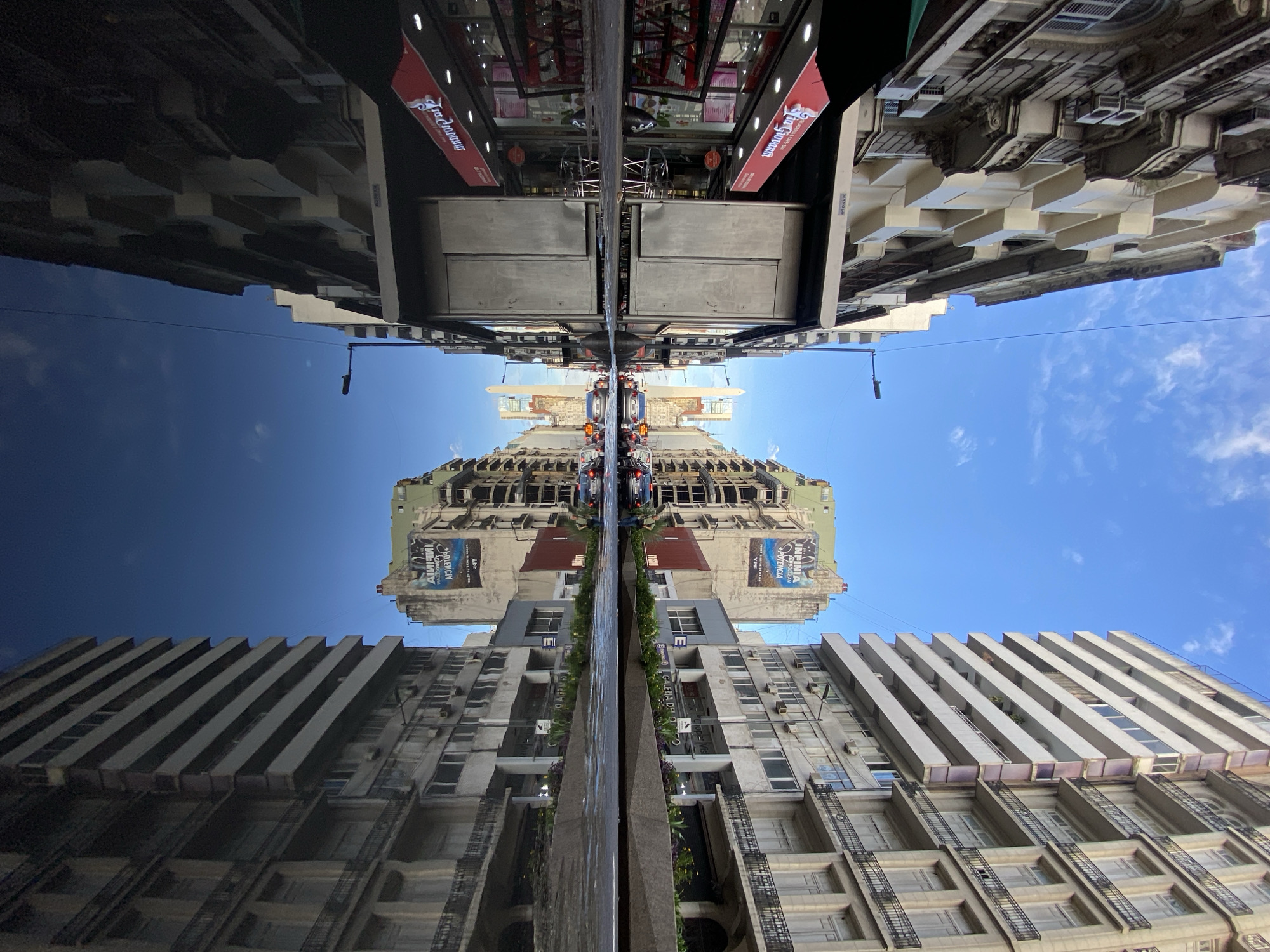 The image depicts a symmetrical view of a street in Buenos Aires, Argentina, seemingly reflected in a wet or polished surface, creating a mirrored effect. Buildings line both sides of the street, varying in height and architectural style, from older ornate structures to more modern, functional designs. Advertisements and signs are visible on some of the buildings, and the sky above is clear with scattered clouds. The mirrored effect creates an abstract composition where the buildings appear to converge at the center.