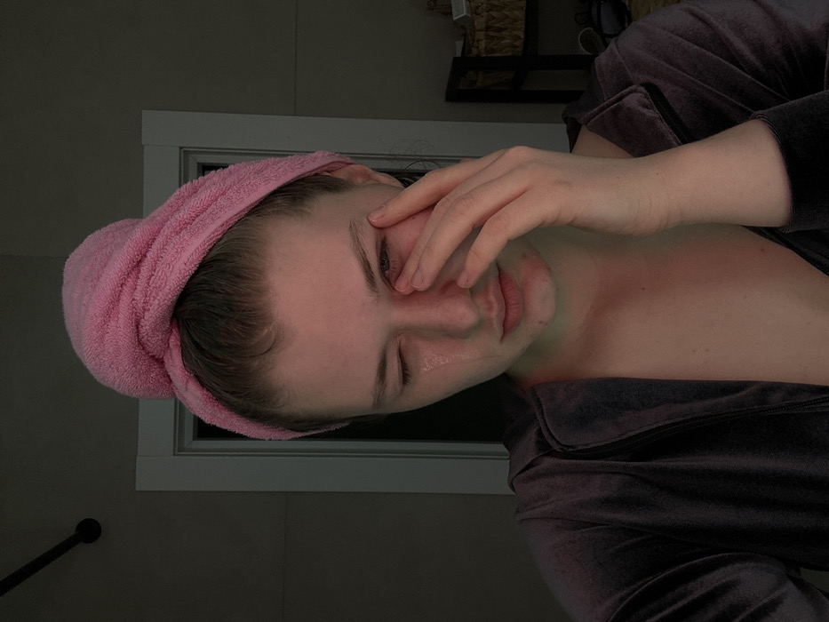 A woman in a purple top and pink towel wrapped around her head rests her head and face on her left hand. Tears are visible on her face. Behind her is a dimly lit bathroom with a small black shelf and a white framed window. The light in the room is low and soft.