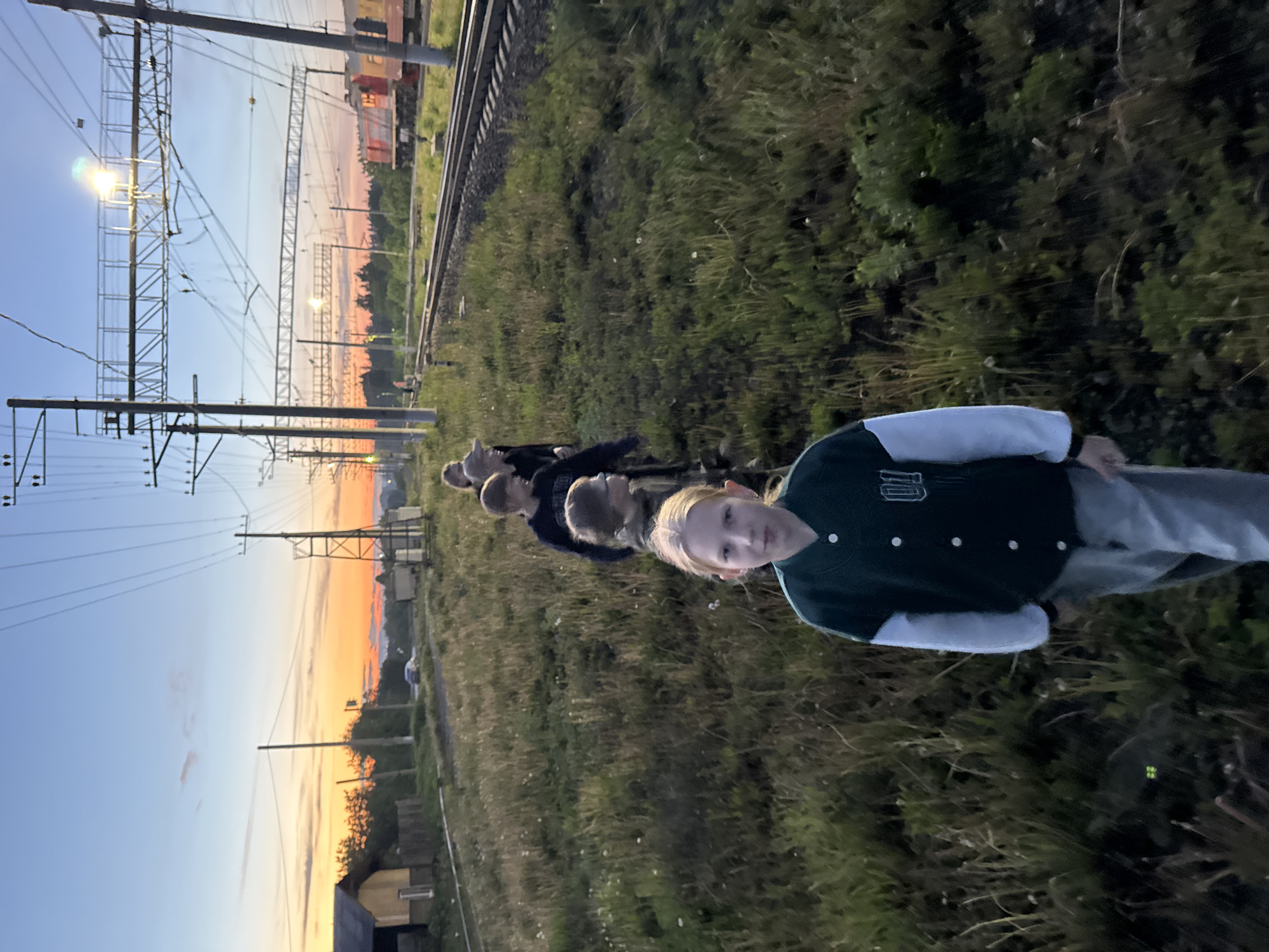 The photograph, taken in Ферма, Russia, depicts a row of five individuals aligned along a railway track. The person closest to the camera is a fair-haired individual wearing a varsity-style jacket. Behind them, the other individuals are partially obscured, facing the same direction. The setting is outdoors, adjacent to a railway line with overhead power lines and infrastructure. The sky shows a transition from blue to orange, suggesting either sunset or sunrise. Vegetation is present in the foreground and along the railway tracks. A building structure is discernible in the distance.