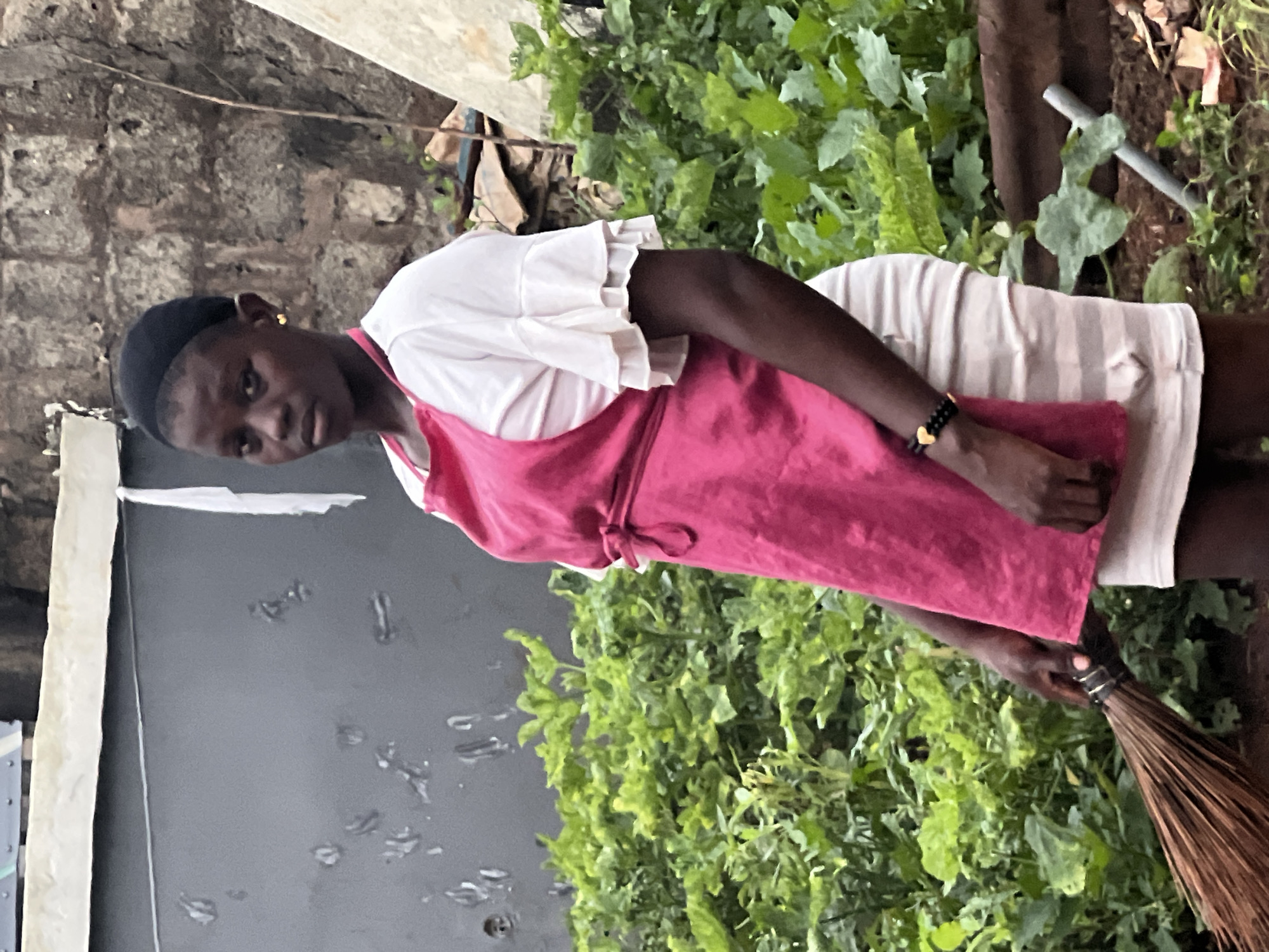 The image depicts a young person, likely a female, standing in what appears to be a garden or a yard. The individual is wearing a pink apron over a white top with ruffled sleeves and a striped skirt or dress. They have a black head covering and are wearing a gold heart-shaped earring and a beaded bracelet on their left wrist. In their right hand, they are holding a broom. The background shows lush green plants, a textured brick wall, and some debris. The setting suggests an outdoor environment, possibly a residential area. The person's gaze is directed towards the camera.