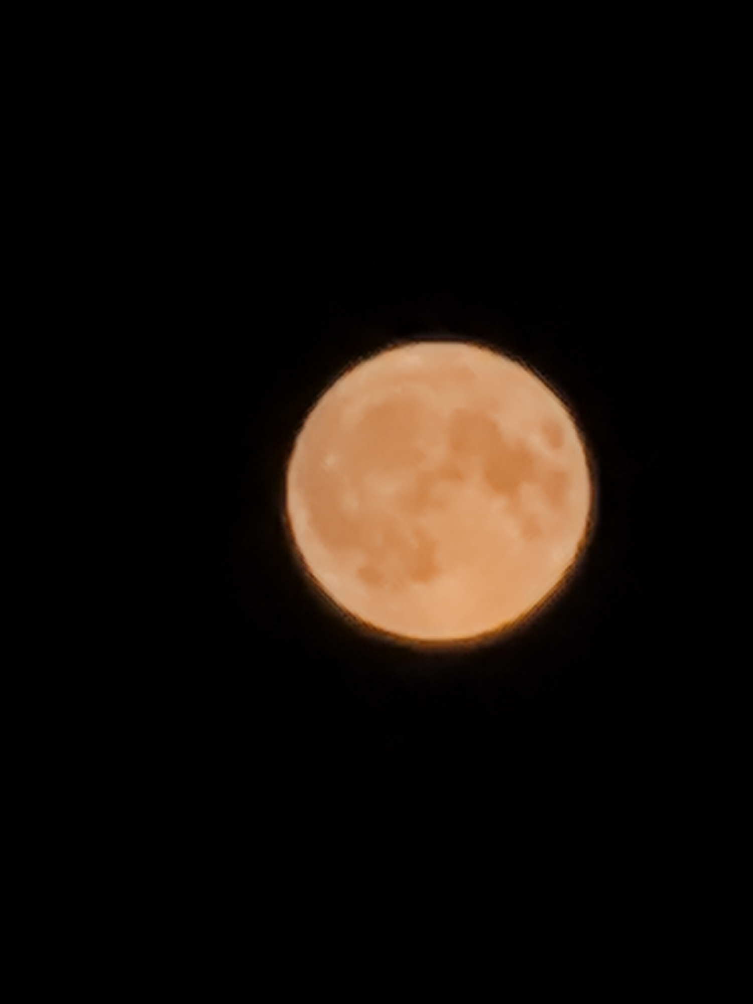 A full, orange moon is depicted against a completely black sky. The moon is centered in the right half of the frame and exhibits some subtle variations in its surface texture, suggesting a slightly uneven topography. The edges of the moon are soft and slightly blurred, hinting at atmospheric conditions or camera focus. There are no other celestial bodies or objects visible in the frame.