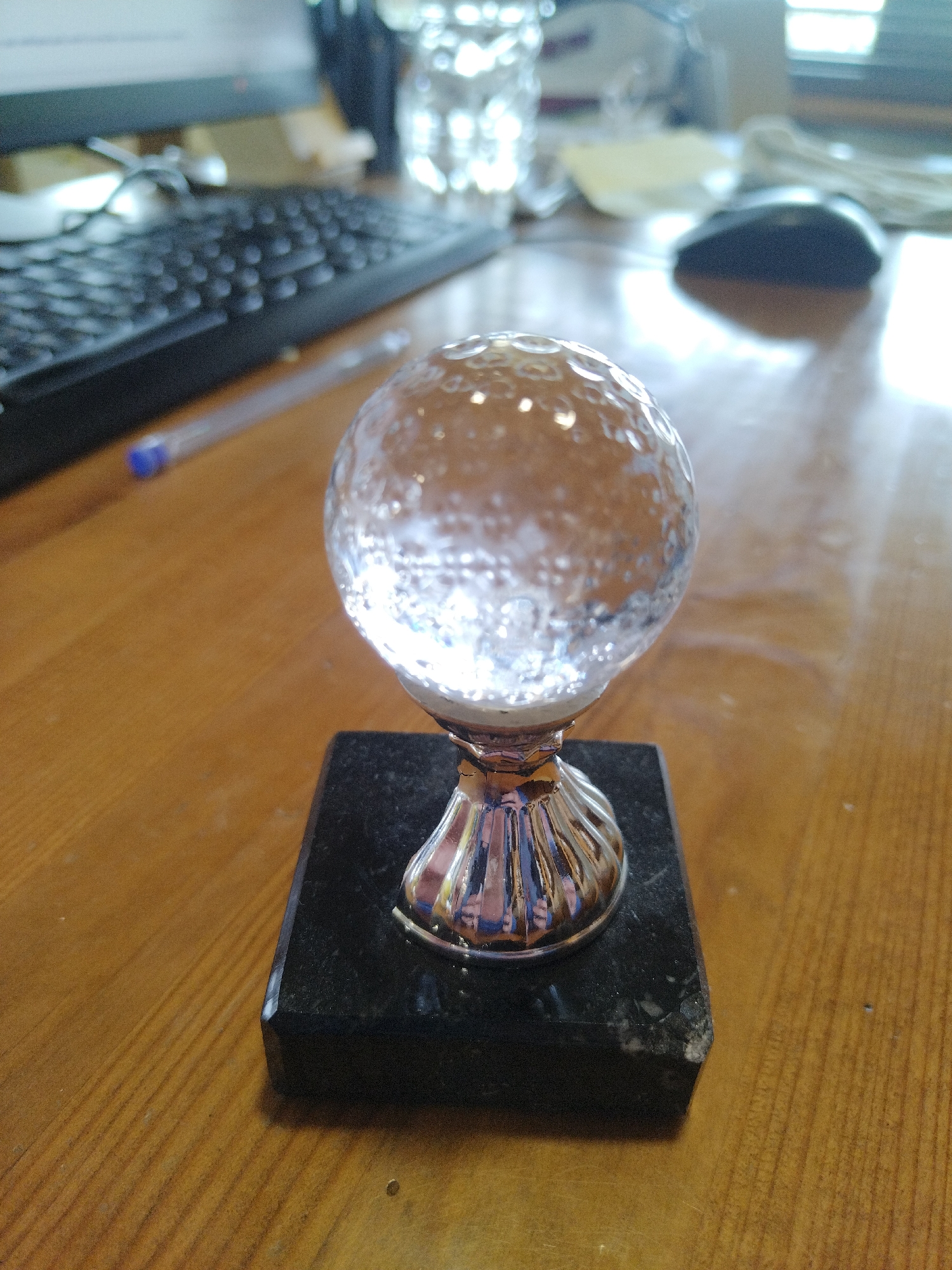 The image shows a crystal golf ball trophy with a shiny, fluted stem sitting on a square, dark-colored base. The trophy is centrally located on a wooden desk or table. In the background, there is a black computer keyboard, a clear plastic pen, a water bottle, a computer monitor, and a black computer mouse. The image does not contain any information that identifies the city of Razlog, Bulgaria.