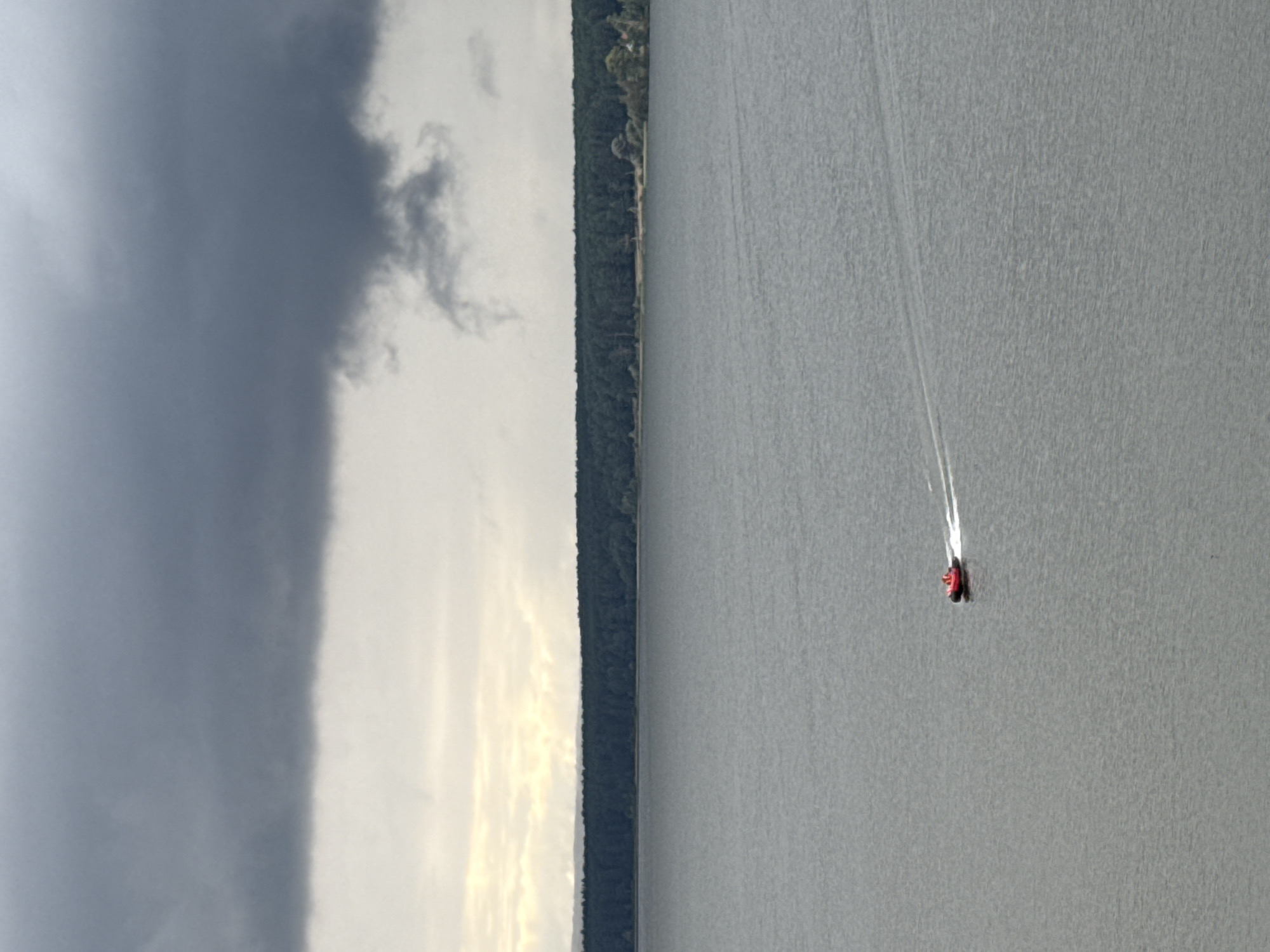 A small, red inflatable boat is moving from left to right across a large body of water. The boat is leaving a white wake behind it. In the distance, a dense, dark green treeline runs horizontally along the horizon. Above the treeline, the sky is overcast with dark, dramatic clouds. The water is calm, with a slight ripple effect. The overall mood of the image is somber and somewhat dramatic due to the stormy sky. This scene appears to be taking place on the Votkinsk Reservoir in Votkinsk, Russia, a large artificial lake known for its scenic beauty and recreational opportunities.