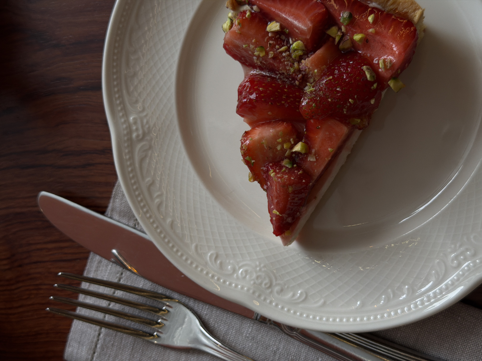 A close-up, high-angle shot shows a slice of strawberry tart on a white, ornate plate. The tart is topped with sliced strawberries and chopped pistachios. To the left of the plate, a fork and knife are resting on a gray napkin. The scene is set against a dark wooden table. The image is focused on the food and cutlery, with the background slightly blurred. This image does not contain information about the location in the city of Saint Petersburg, Russia.