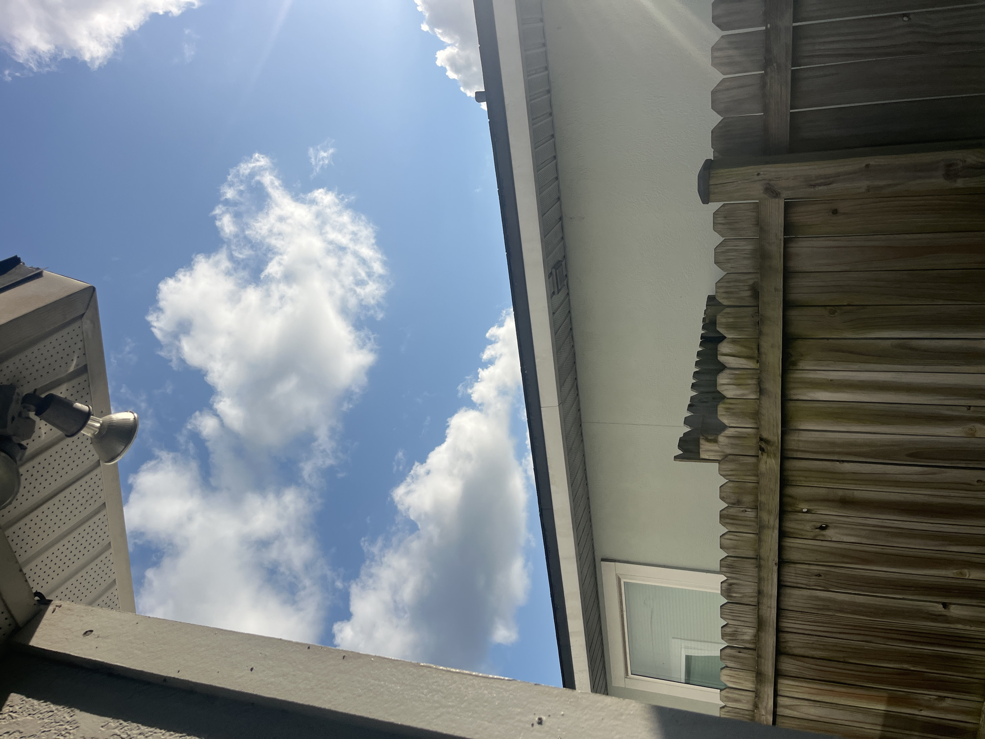 A clear, blue sky with scattered white clouds fills the upper portion of the image. A portion of a gray, textured wall and a white soffit with a light fixture are visible on the left and top of the frame respectively. Below the sky is a light green building with a dark roofline and a white-trimmed window. A wooden fence with vertical planks and a decorative top edge runs across the lower half of the image. The scene appears to be an outdoor residential setting in Jacksonville, Florida, United States of America, likely on a sunny day.