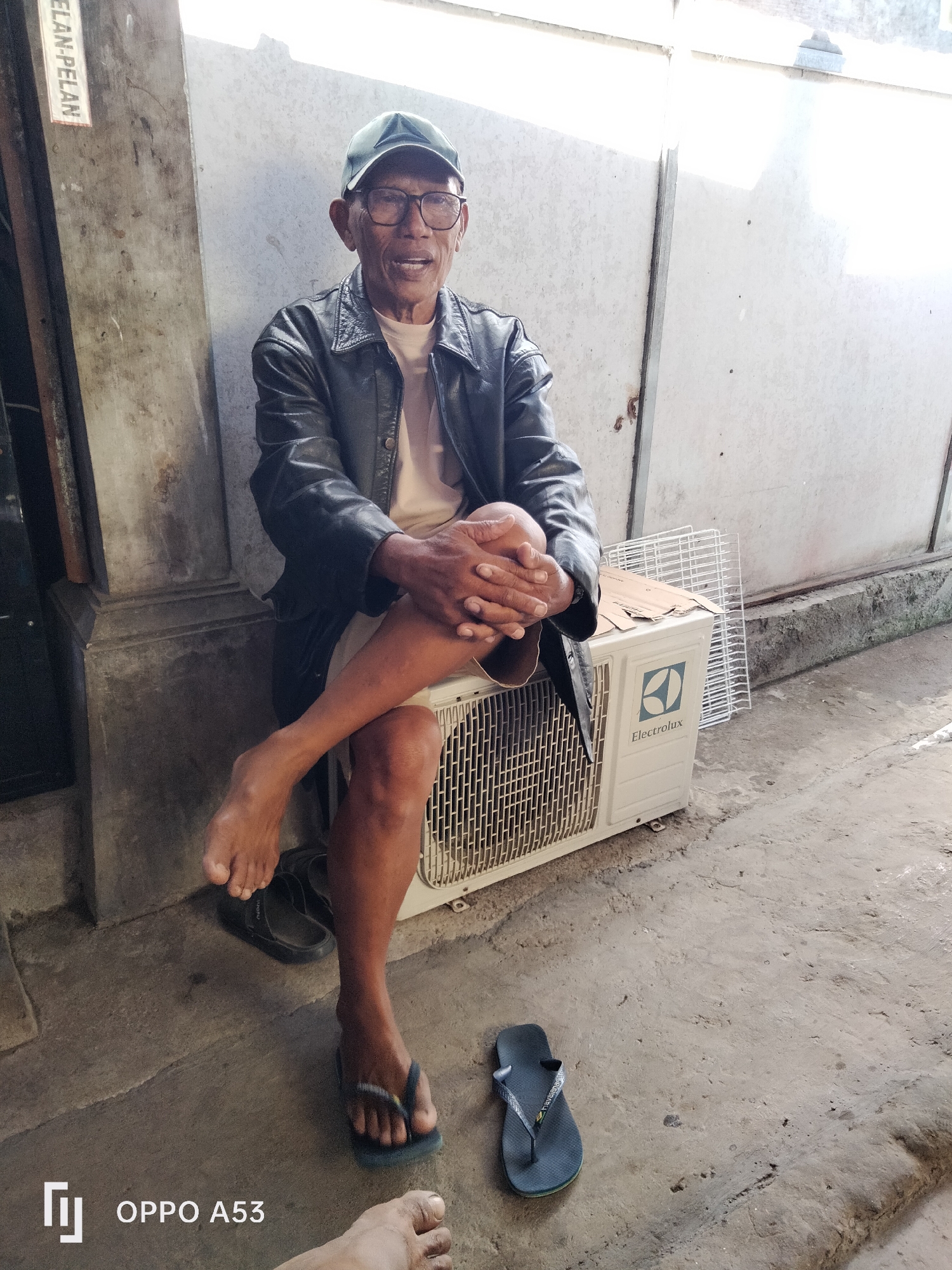An elderly Indonesian man with glasses and a dark baseball cap sits on an outdoor air conditioning unit. He wears a black leather jacket over a light-colored shirt and has his legs crossed, his bare feet visible. One foot rests on a pair of black flip-flops. Another single dark flip-flop is on the ground to his right. The man is sitting on a paved surface in front of a light-colored, corrugated metal wall. To his left, a wooden pillar is visible with a sign that has "ELAN-PELAN" written on it. The sun is shining brightly, casting shadows. The overall scene appears to be an outdoor area in the city of Denpasar, Indonesia.