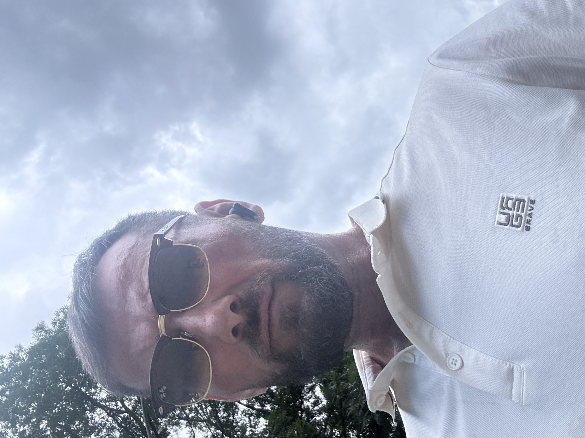 A man is looking upwards at the camera, with trees and a cloudy sky in the background. He has short, dark hair, a beard, and is wearing sunglasses and a white polo shirt. The man is in the foreground, and the background is out of focus. The image appears to be a selfie taken outdoors, likely in a park or natural setting.