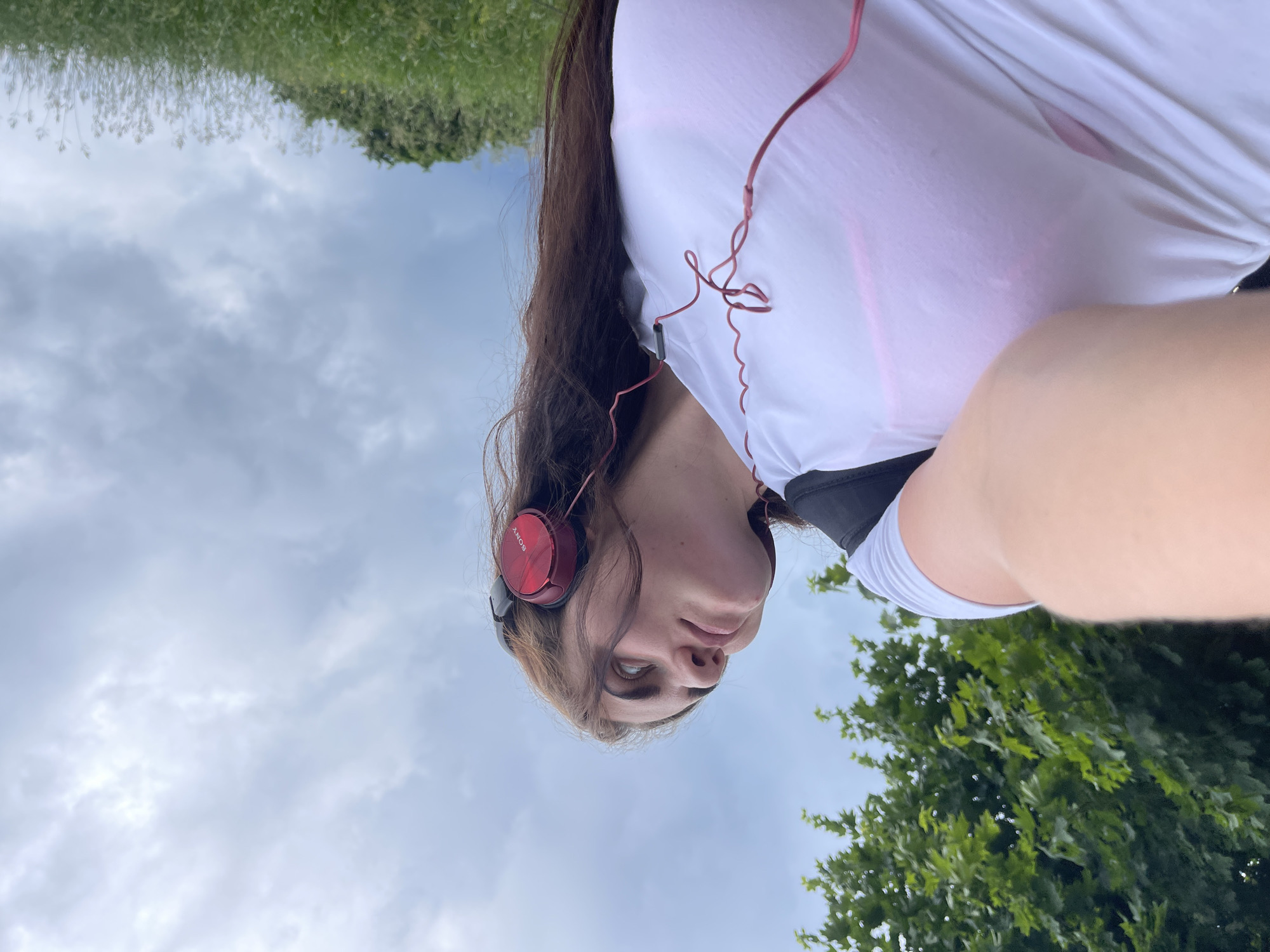 An outdoor, low-angle shot shows a young woman with long, dark hair wearing red Sony headphones. She is looking up and to her left. She is wearing a white t-shirt with a pink sports bra underneath, and a black backpack strap is visible. The background consists of a cloudy sky and green foliage, with trees on the left and a grassy slope on the right. This scene is located in Ostrov, Russia.
