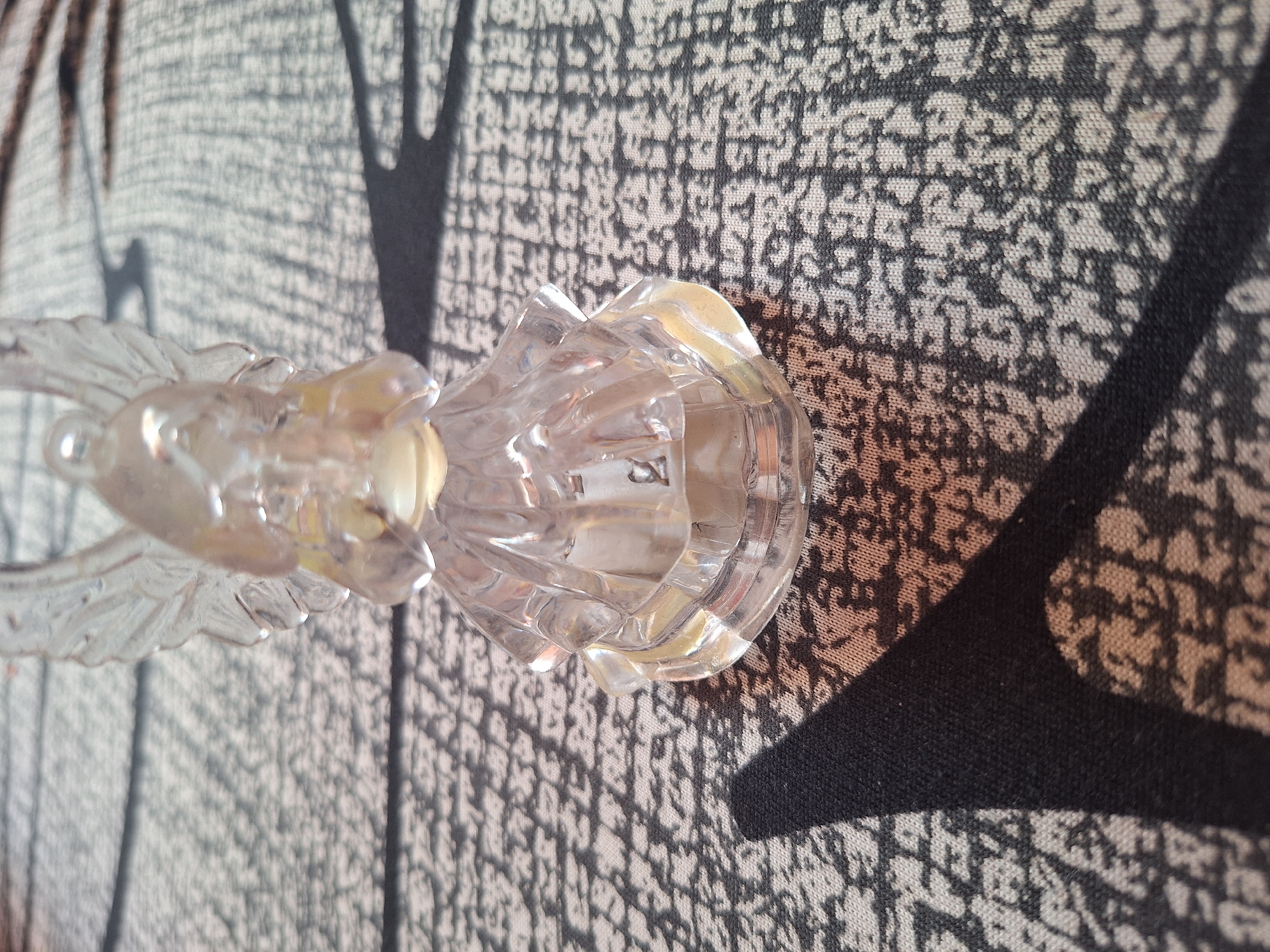 A clear, plastic angel figurine is positioned on a patterned surface, casting distinct shadows. The figurine is translucent, with delicate wings and a detailed dress. The surface beneath it has a repeating, abstract pattern in dark grey and white. Sunlight creates sharp, dark shadows from the figurine and its surroundings, indicating an outdoor or well-lit indoor environment. No specific location in Varna, Bulgaria is discernible from the image.