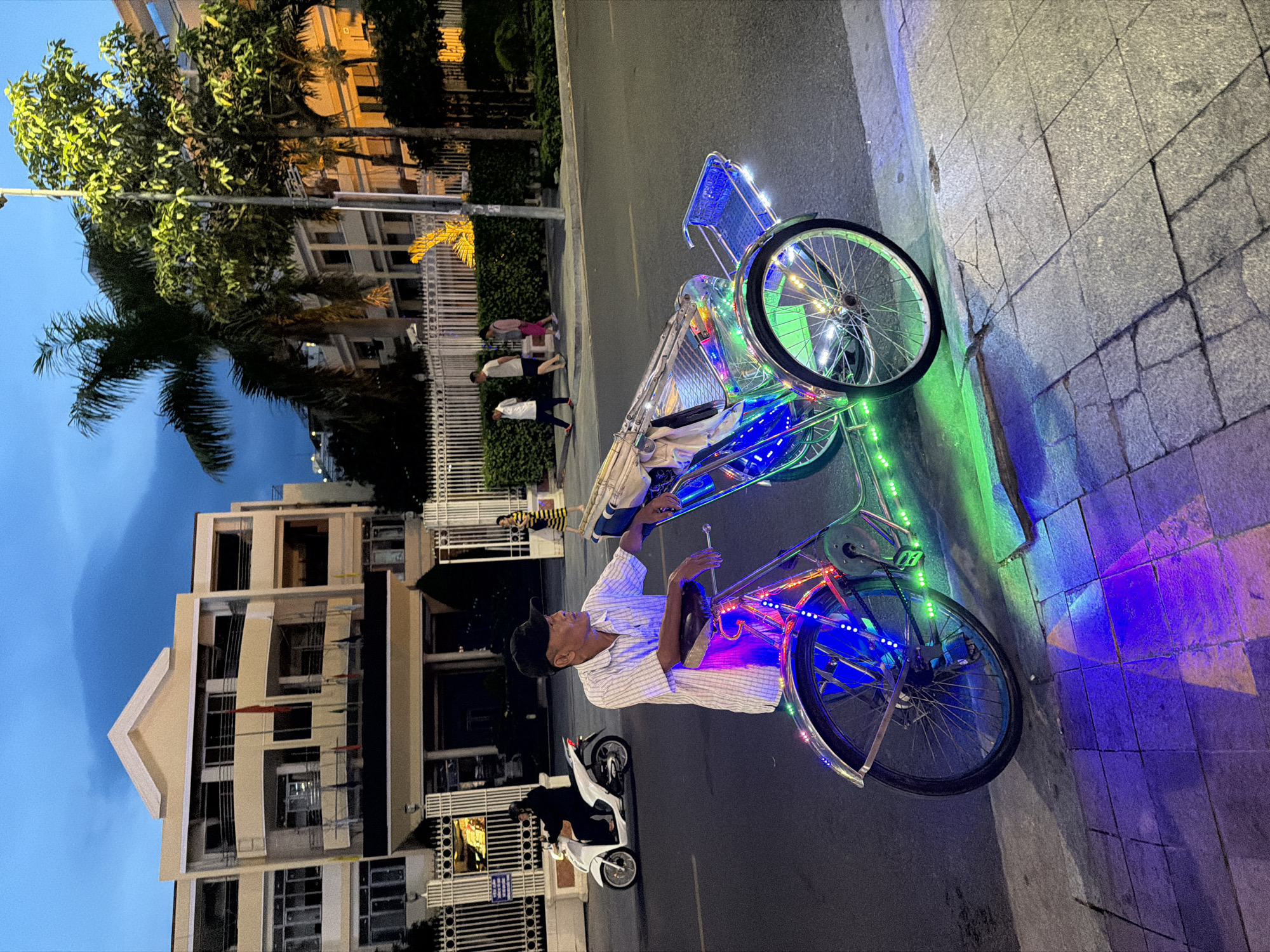 This image depicts a street scene in Nha Trang Ward, Vietnam, likely in the evening or at night, given the artificial lighting. The primary focus is on a decorated pedicab, adorned with bright blue and green LED lights that cast colorful reflections on the pavement. A man, wearing a checkered shirt and a dark cap, sits in the driver's position of the pedicab, appearing to be engaged in conversation or waiting for passengers.
In the background, a modern building with multiple floors and balconies is visible. The architecture suggests an urban setting. Several individuals are seen walking on the sidewalk near a white fence. Further down the street, a white scooter is parked, with a person in dark clothing seated on it. The overall atmosphere suggests a typical street scene in a Vietnamese city, with a blend of traditional transportation and modern infrastructure, illuminated by festive lights. The time of day appears to be dusk or nighttime, as indicated by the illuminated lights and the twilight sky.