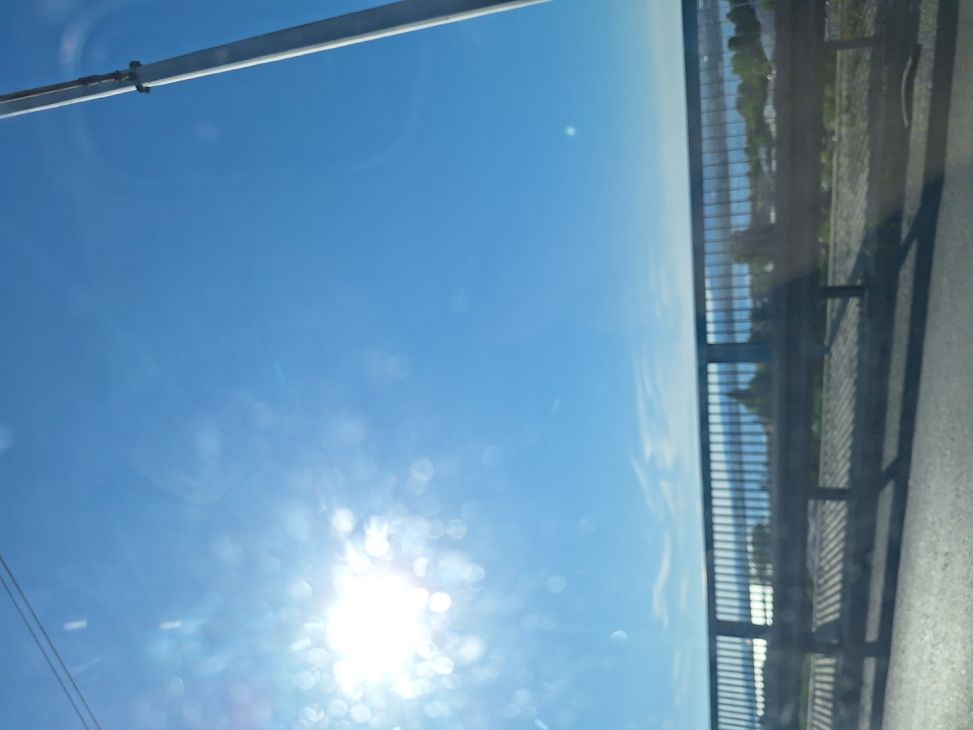 The image captures a bright, sunny day with a clear blue sky. The sun is prominently featured on the left side of the frame, casting a glare that creates bokeh effects. A tall, grey pole, possibly a lamppost, stands on the right side. In the middle ground, a metal railing stretches horizontally across the scene, suggesting a bridge or an elevated walkway. Beyond the railing, a body of water is visible, with a distant shoreline dotted with trees and buildings. The foreground shows a road surface, indicating the image was likely taken from a moving vehicle. The overall impression is a scenic view, possibly from a bridge overlooking a bay or lake within the city of Varna, Bulgaria, given the characteristic blue sky and water views common in coastal Bulgarian locations. There are no people or animals visible in the image.