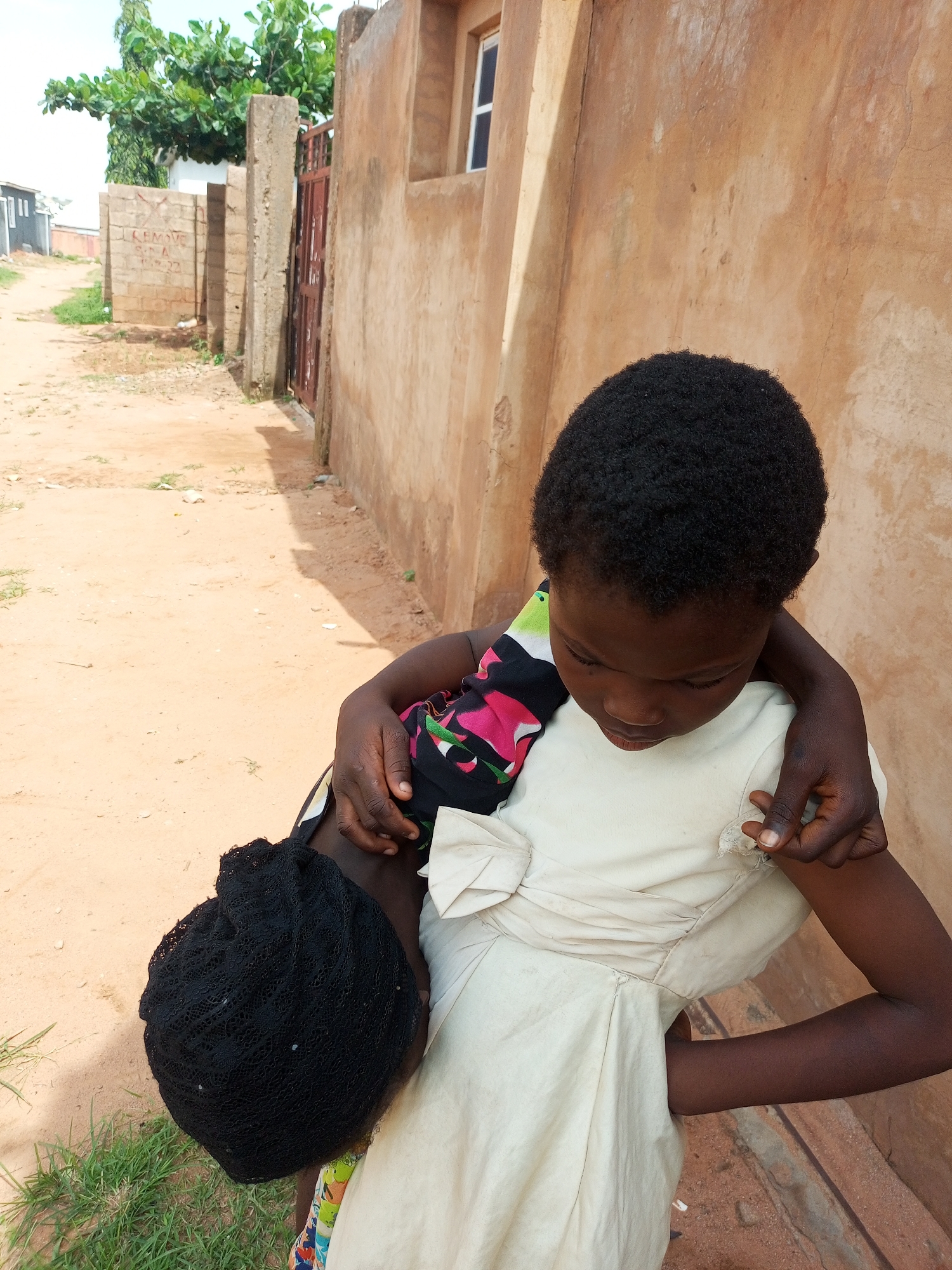 A young girl with dark, curly hair and a light-colored dress embraces another child who is wearing a black head covering. The older girl's arm is around the younger child's shoulder and her face is turned downwards, looking at the other child. The younger child's head is bent forward, obscuring their face. They are standing on a dirt path next to a textured, ochre-colored wall, with a hint of green grass to the left. In the background, there is a metal gate and a concrete structure, suggesting an outdoor environment, possibly in Gombe, Nigeria. The overall impression is one of an intimate and gentle interaction between two children.