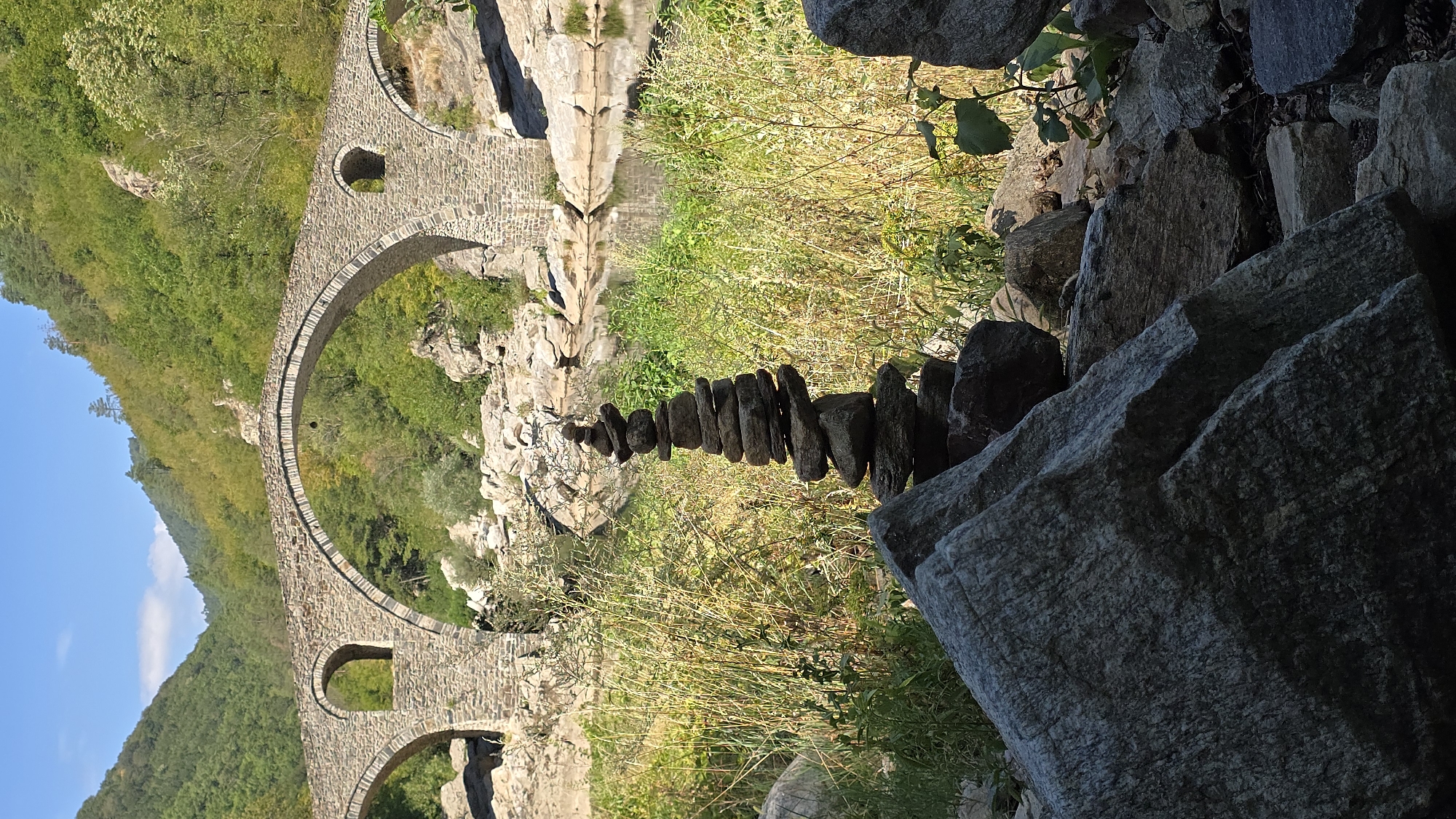 The image shows the Devil's Bridge, an ancient Roman bridge located in the Rhodope Mountains, near the village of Dyakovo, Ardino Municipality, Kardzhali Province, Bulgaria. The bridge spans a gorge over the Arda River. In the foreground, a cairn of stacked stones is positioned in front of lush green vegetation and tall grass. The stone bridge, constructed of massive stones, features two large arches and a smaller arched opening in the parapet. The surrounding environment consists of rocky terrain and dense green forestation covering the steep hillsides. The sky is clear blue with a few wispy clouds. The lighting suggests it is daytime, likely late morning or early afternoon, casting shadows and highlighting the textures of the stone and vegetation. No people are visible in the scene.
