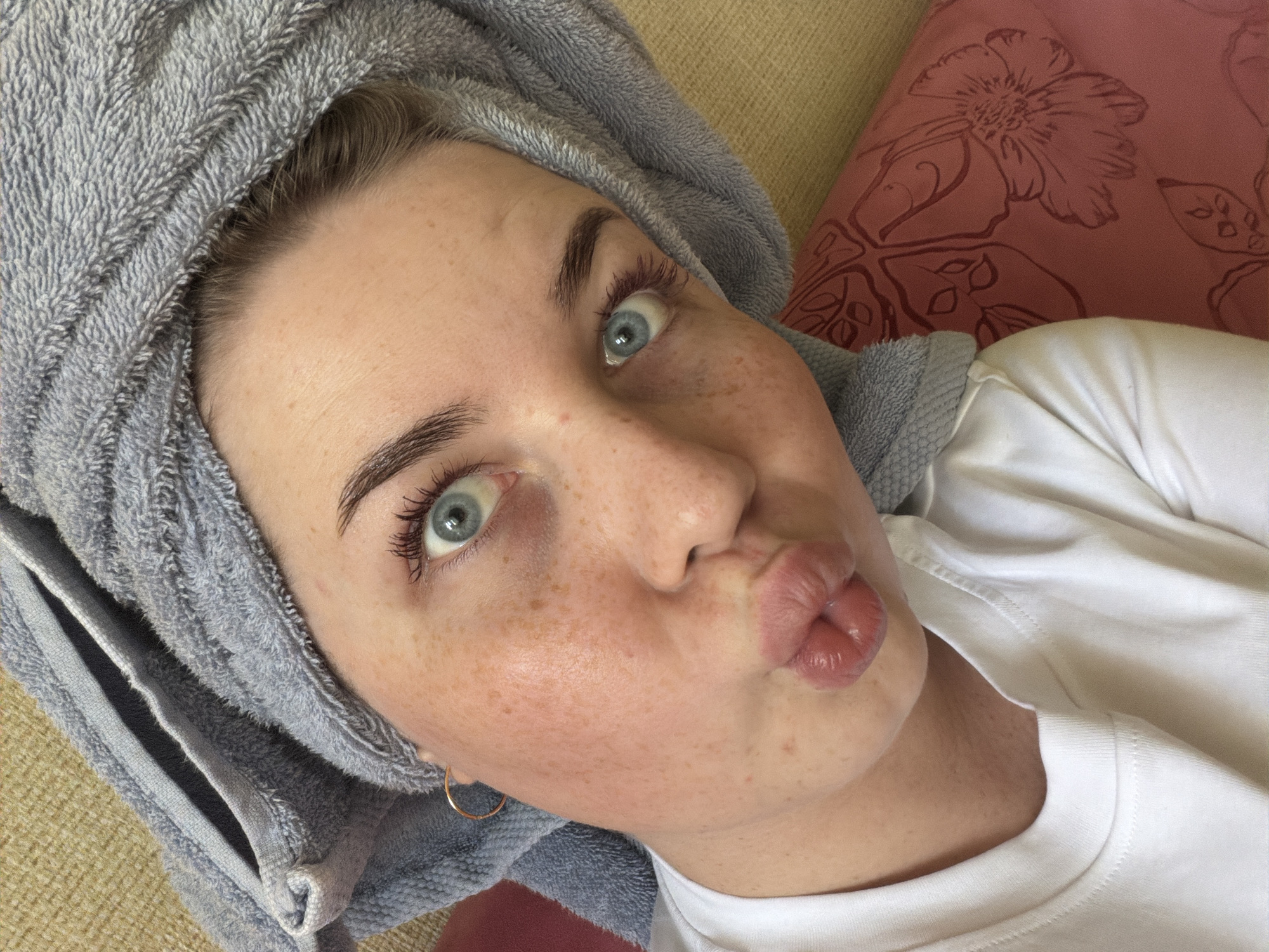 A fair-skinned woman with freckles and light blue eyes poses for a selfie while puckering her lips. Her hair is wrapped in a gray towel. She is wearing a white shirt and a gold hoop earring is visible. A pillow with a floral pattern is in the background. There is no information to determine if this image was taken in Chaikovsky, Russia.