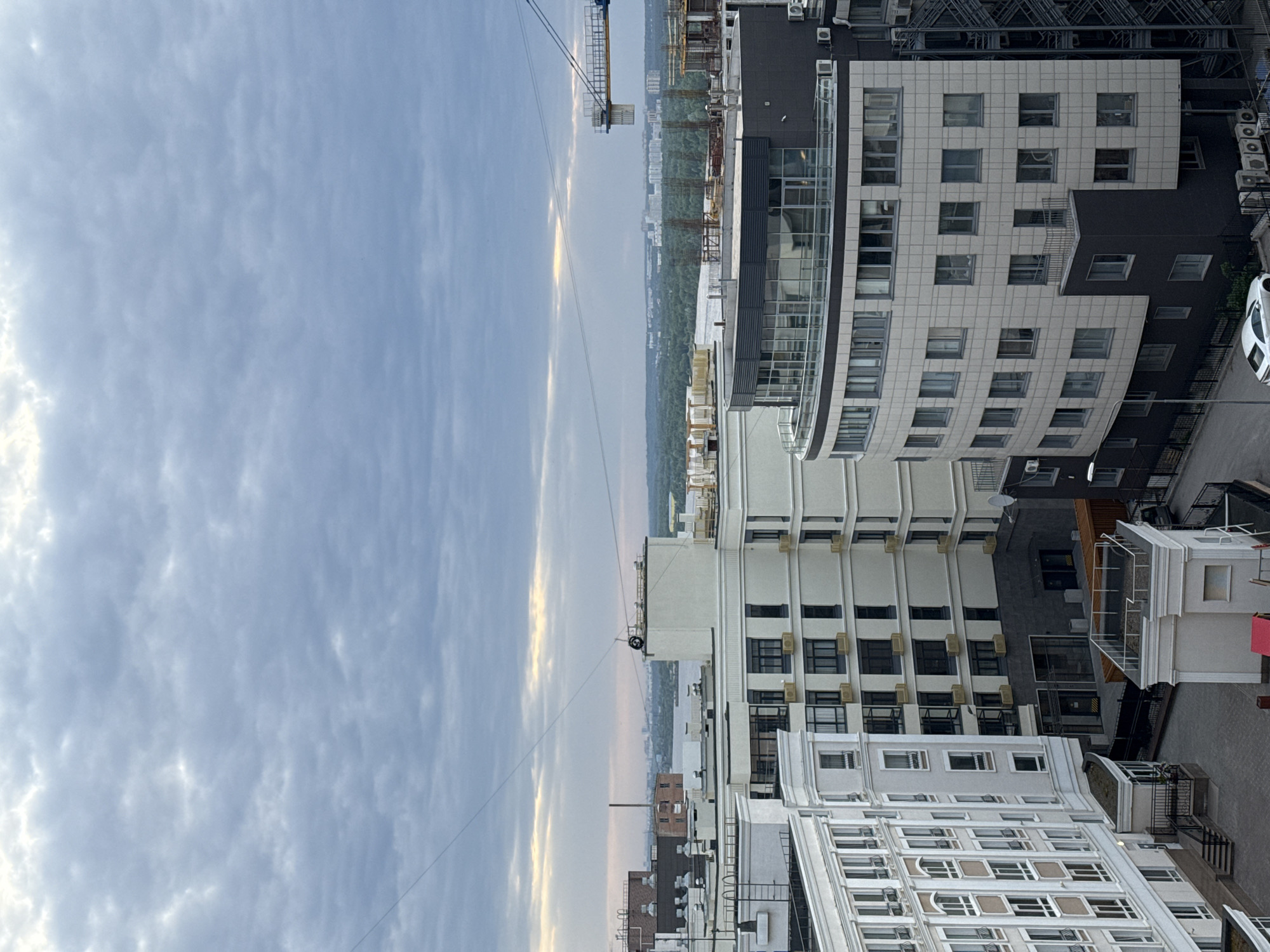 The image shows an urban scene with buildings under a cloudy sky. The architecture varies, with some buildings displaying modern designs and others more classic features. A construction crane is visible in the distance, indicating ongoing development. The image is taken from an elevated perspective, likely from a high floor of a building. The location is the city of Perm, Russia. The environment is characteristic of a city, including dense buildings and limited greenery. There are buildings with air conditioning units installed. There is a parked white car next to one of the buildings.