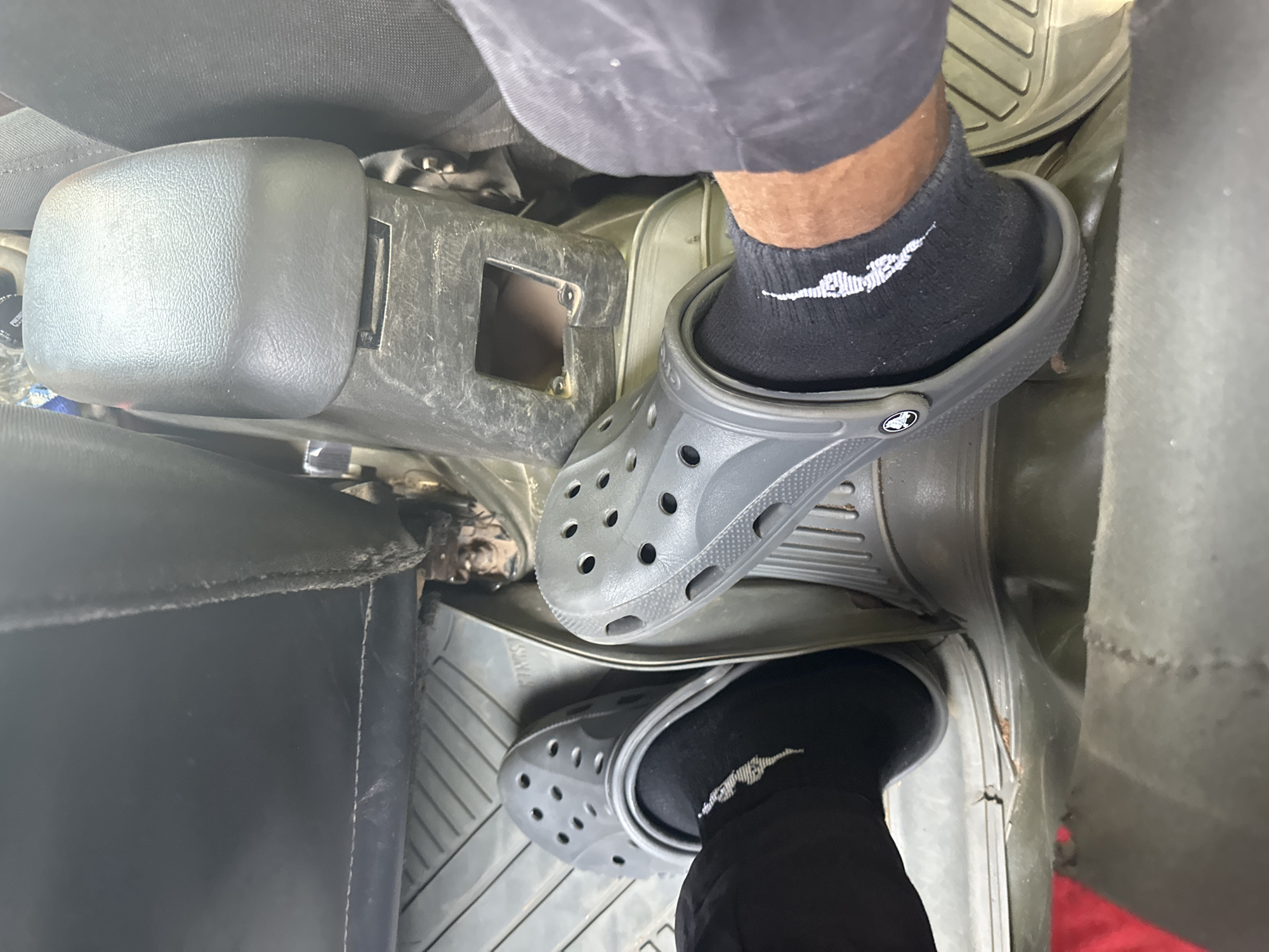 The image depicts the interior of a vehicle, focusing on the feet of a person wearing black socks and grey Crocs. The vehicle appears to be a utility vehicle or truck, with a rugged interior featuring rubber floor mats and a prominent grey plastic center console. The floor mats are ridged and somewhat dirty, showing signs of wear. The center console has a rectangular opening and appears dusty. The seats are visible, covered in dark fabric, with the edge of a seat in the foreground. The overall environment suggests a practical, utilitarian vehicle, possibly in a dusty or unpaved area, consistent with some locations within Abuja, Nigeria. The lighting is natural, indicating daytime. There are no visible landmarks or specific indicators to pinpoint the exact location within Abuja.