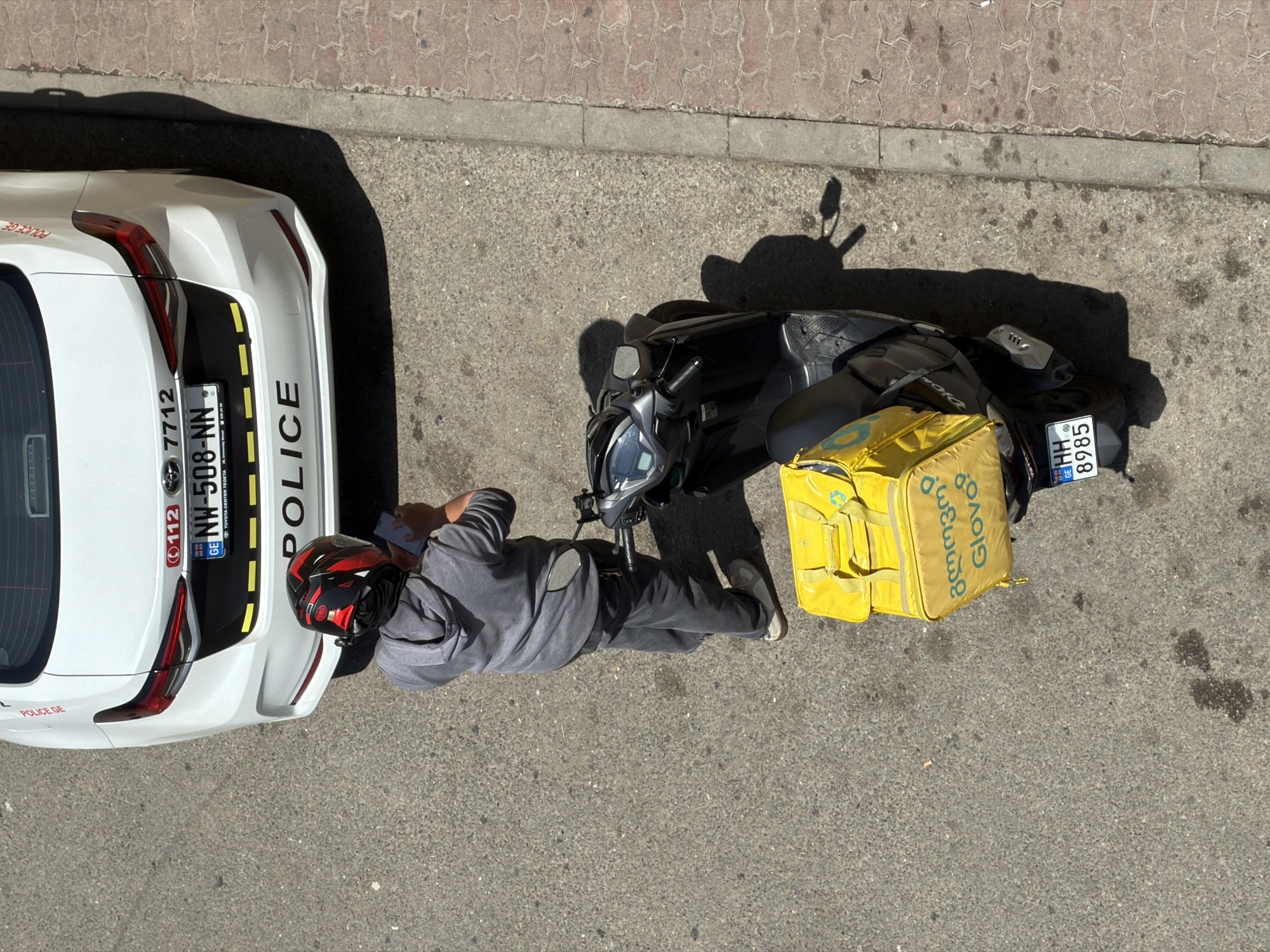 The scene is an aerial view on a sunny day, showing a man in a grey sweatshirt, grey pants, and a red helmet standing next to a white police car and a black motorcycle. The police car, partially visible on the left, bears the markings "POLICE GE" and "NW-508-NN". The motorcycle has a yellow Glovo delivery bag attached to it and a license plate reading "HH 8985". The image indicates the location is in Tbilisi, Georgia, given the Georgian writing and license plates.