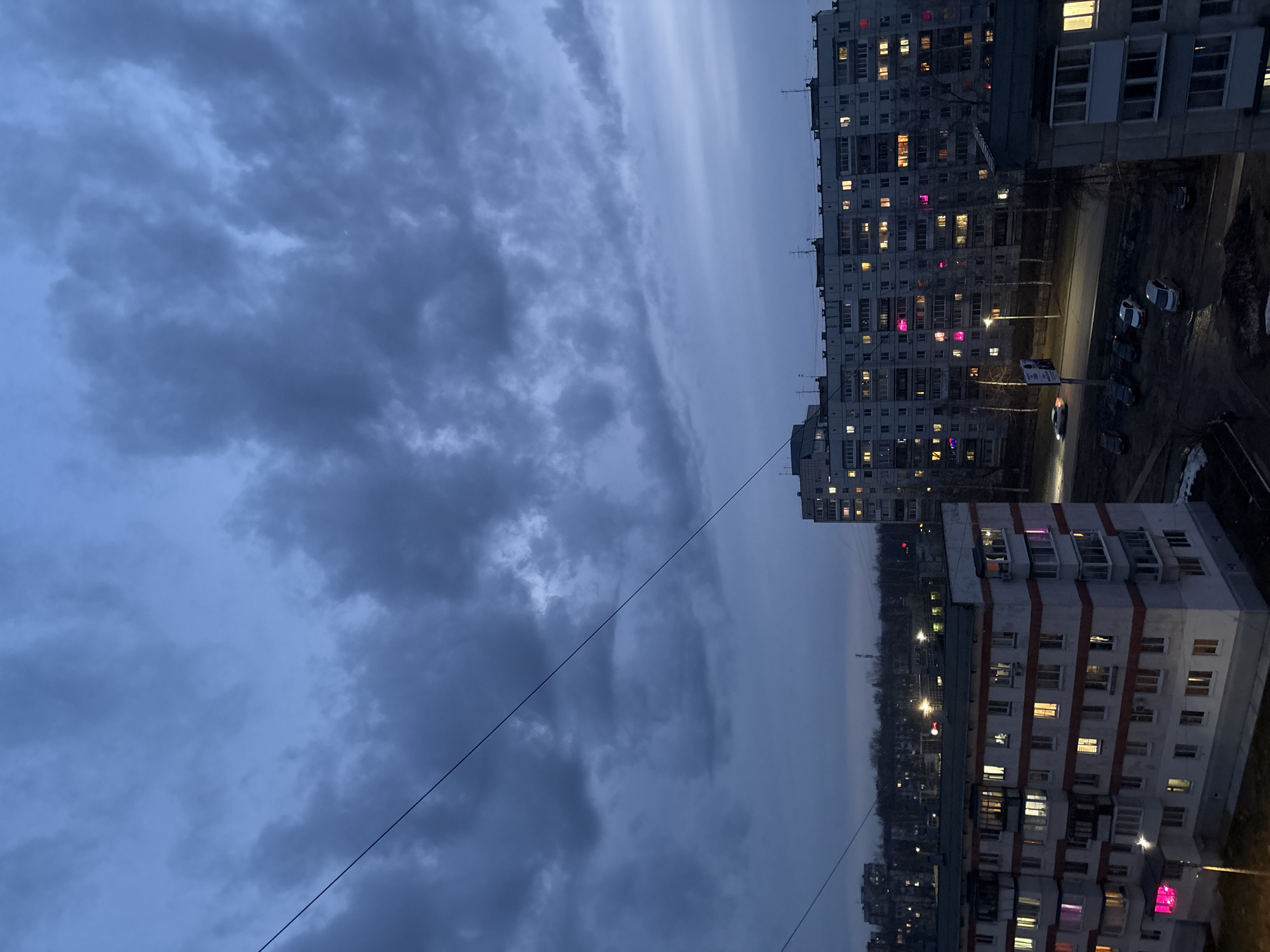 The image depicts an urban scene in what is likely Chaikovsky, Russia, based on the style of architecture. The photograph is taken from a high vantage point, possibly an upper floor of a building, looking out over other apartment buildings and a heavily clouded sky.
The right side of the image contains several multi-story apartment buildings with numerous windows. Many windows are illuminated, suggesting it is evening or night. Some windows display colors, such as pink. Parked cars are visible next to the apartment buildings.
The left side of the image is dominated by a dark, cloudy sky. Power lines extend across the frame.