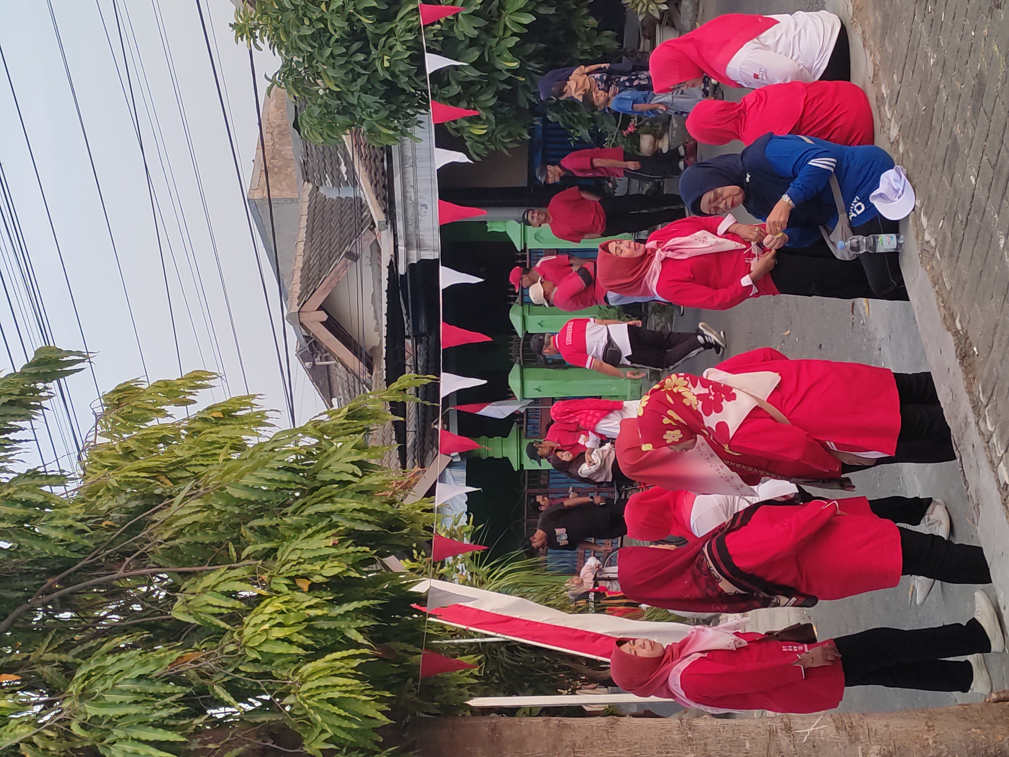 In Ponorogo, Indonesia, an outdoor scene unfolds with a group of people gathered on a street, adorned with red and white festive decorations, including triangular pennants strung across the scene and a vertical red and white banner. The majority of individuals are dressed in red tops, with many women wearing hijabs in various shades of red and pink. A few are in black pants and white sneakers, while others are in white tops with red accents.
In the foreground, several women are standing and conversing. One woman in a red hijab and pink scarf looks to the left, while another beside her, also in red, faces away from the camera. To their right, a woman in a bright red hijab and a patterned scarf is seen from the side. Further to the right, a woman in a blue hijab and track jacket is seated on a curb, appearing to be preparing a small item in her hands. A water bottle and a white cap are on the ground beside her.
In the middle ground, a group of men and women are also present. Some men are in red shirts and caps, while others are in black. A man in a black t-shirt stands near a bicycle. A woman in a red outfit holds a child. Several other individuals in red are interacting with each other.
The background shows residential houses with tiled roofs and a tree with long, green leaves partially obscuring the view. Overhead, electrical wires crisscross the sky. The lighting suggests it is daytime, likely late afternoon or early morning due to the soft light. The overall atmosphere appears to be one of a community gathering or celebration.