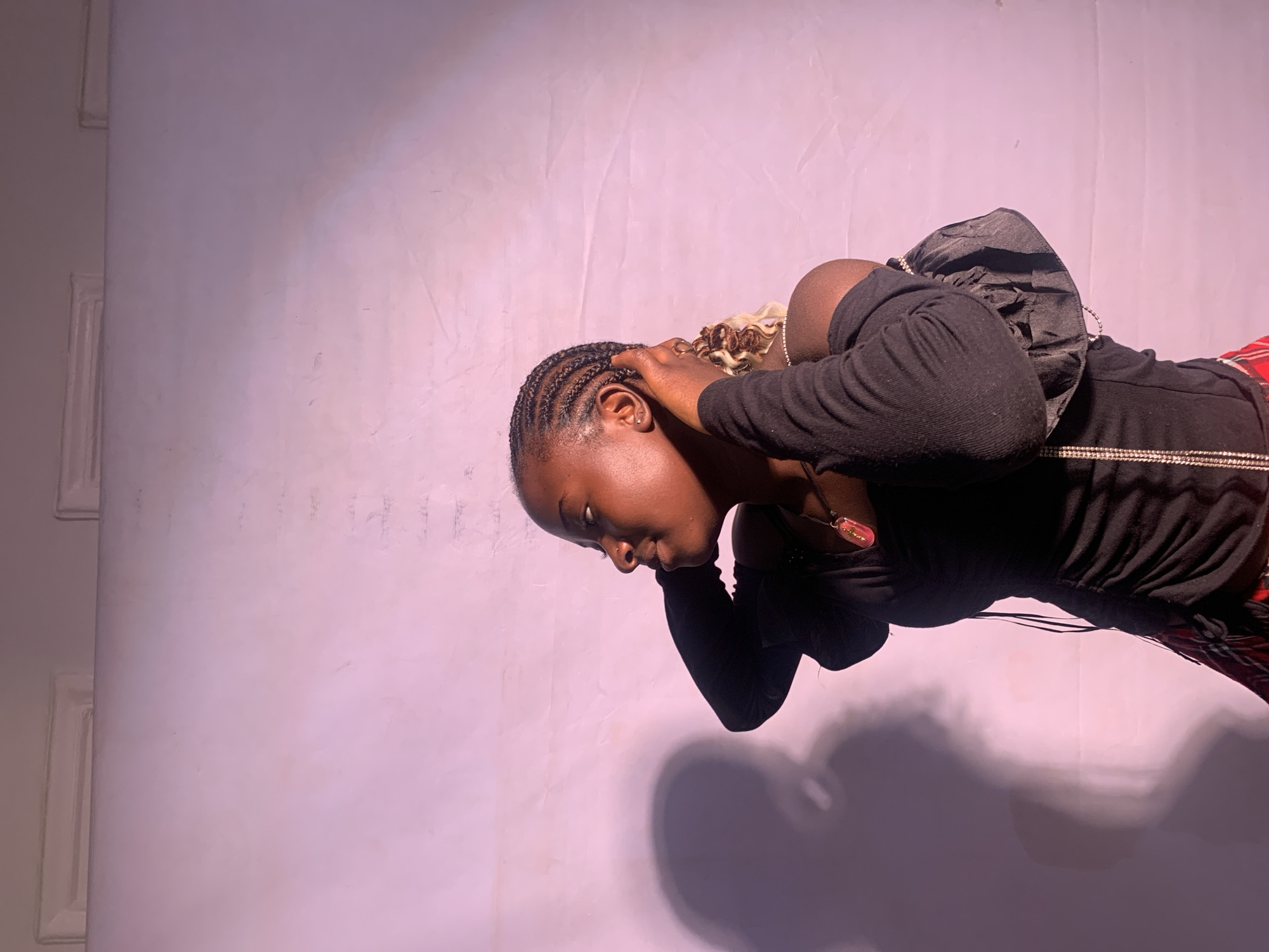The image depicts a young, dark-skinned woman with braided hair and some blonde highlights, wearing a black long-sleeved shirt and red plaid pants. She has a black backpack slung over her shoulder. She's turned away from the camera with her hands near her head. Her shadow is cast on a pale purple background. The location is possibly in Jos, Nigeria, based on the prompt's instructions, but there are no features definitively linking the image to this location. The scene appears to be a portrait-style photograph taken indoors.