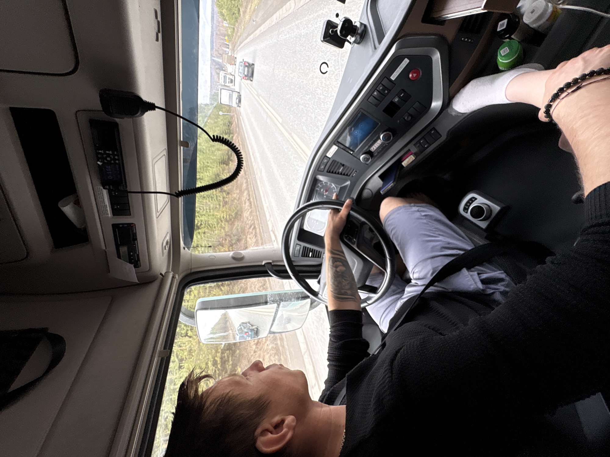A person is driving a truck on a paved road with lane markings. Several other vehicles are visible ahead in the distance. The driver is wearing a black long-sleeved shirt, light-colored pants, and a white sock. The truck's interior features a steering wheel, dashboard with controls, a radio, and a microphone. The side mirror is visible showing the road behind. The location is inferred from the other vehicles on the road as somewhere in Iiul'skoe, Russia. The environment visible through the windshield and side mirror includes trees alongside the road. The driver is holding the steering wheel, presumably driving.