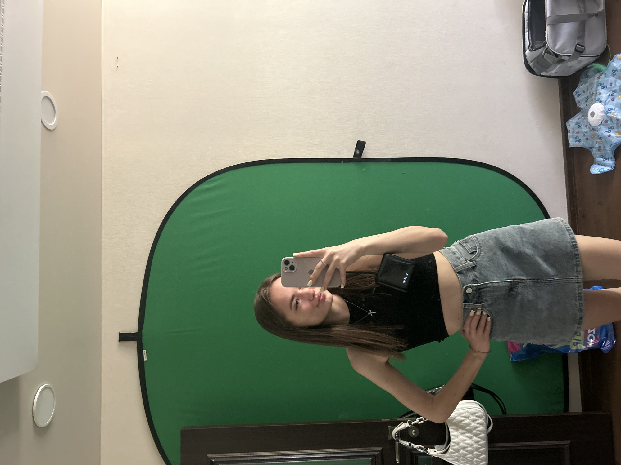 A young female, appearing to be in her late teens or early twenties, takes a mirror selfie in an indoor room in the city of Unknown, Unknown. She has long, straight, light brown hair and is wearing a black sleeveless crop top, a light-colored denim mini-skirt, a silver cross necklace, and a ring on her left hand. Her fingernails are painted a light yellow or beige. She holds a light-colored iPhone with multiple camera lenses in her right hand, which is visible in the reflection. Her left hand rests on her hip.
Behind the female is a large, freestanding, oval-shaped green screen (chromakey backdrop) with a black border, occupying a significant portion of the background. To the left of the green screen, a dark brown door with a silver handle is partially visible. A white quilted handbag with a silver chain strap hangs from the door handle.
The room features beige or off-white walls and a white ceiling with two circular recessed lights. The floor is dark wood or laminate. On the floor, to the left of the female, a crumpled blue plastic bag with white text is present. To the right of the female, on the floor, there is a light blue patterned baby playmat with stars and other designs, featuring a white circular module in the center. Adjacent to the playmat is an open grey soft-sided travel bag or pet carrier with mesh sides.