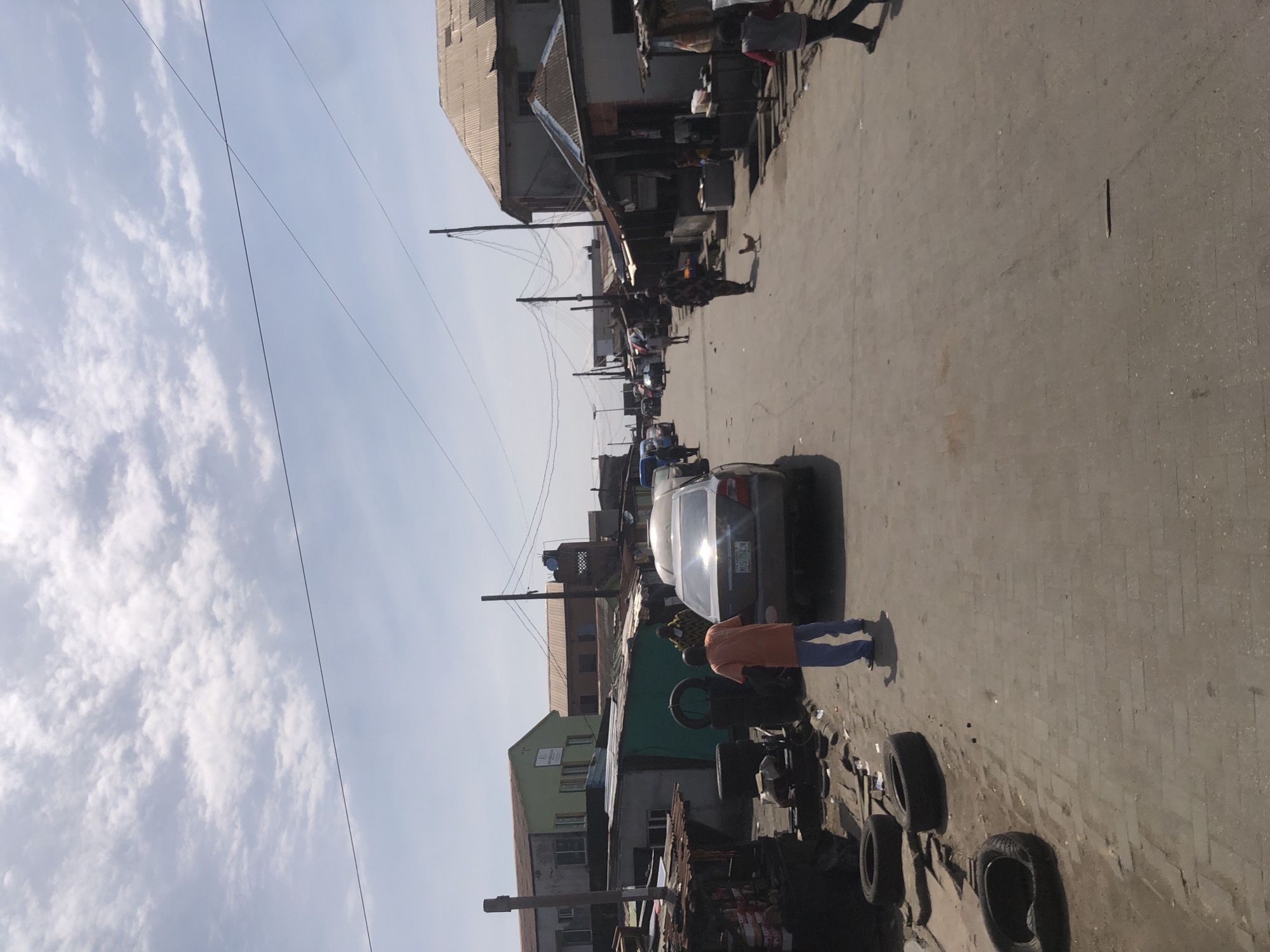 The image depicts a street scene in Ado, Nigeria, under a bright, clear sky with scattered clouds. The perspective is from above, looking down onto a paved street lined with buildings and various objects.
Several people are visible in the scene. In the foreground, a person in an orange shirt and blue pants is standing, facing away from the camera, near a piece of machinery with large wheels. Further down the street, more individuals are walking or standing, some appearing to be interacting. Various vehicles are parked or driving along the street, including a silver car with tinted windows and a license plate that reads "NH5 704 RN". Other cars and what appears to be a motorcycle are also present.
The buildings lining the street are varied in construction, with some appearing to be shops or residences. They are a mix of materials, including corrugated metal roofs and painted walls, suggesting a bustling urban environment. The street itself is paved with interlocking bricks or stones, and there are various items scattered along the sides, including tires, debris, and what looks like market stalls. Overhead, power lines crisscross the sky, a common feature in many urban settings.
The time of day appears to be morning or afternoon, indicated by the strong sunlight casting shadows. The overall impression is one of everyday life in a busy Nigerian town, with people going about their daily activities amidst the urban infrastructure.
