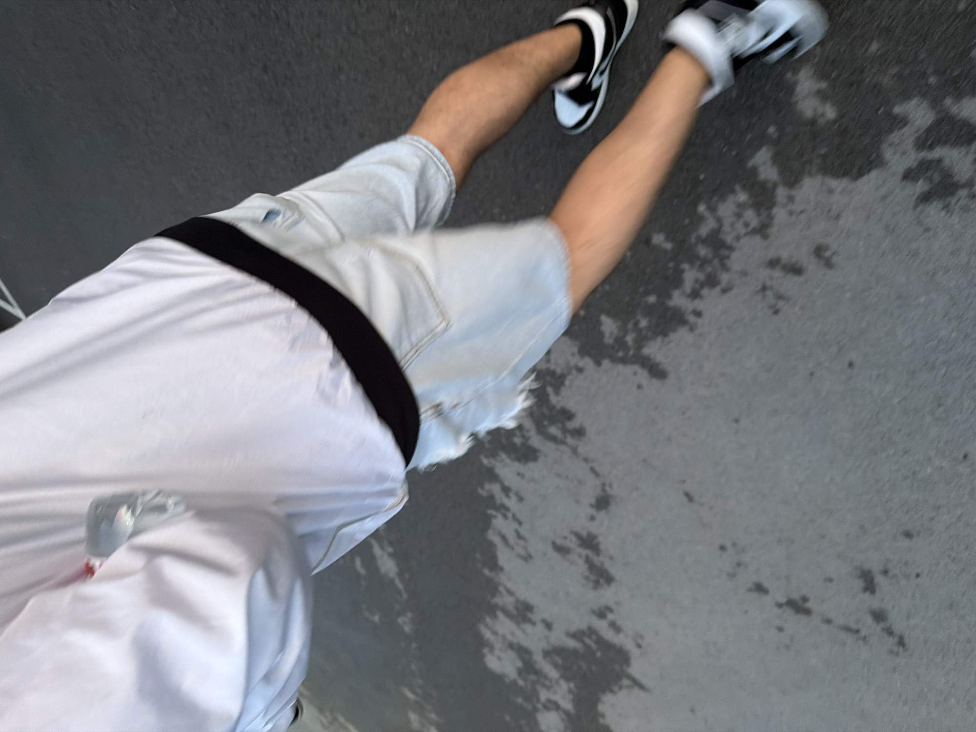 The image presents a top-down, tilted view of a person in motion, captured from the waist down. The subject is wearing a white short-sleeved top, light blue denim shorts, and black and white athletic sneakers. A black strap, possibly from a bag or fanny pack, is visible across the person's back. Motion blur is evident on the legs and shoes, indicating the person is walking or moving at a brisk pace.
The scene is an outdoor paved area, likely a street, sidewalk, or concrete path, consistent with an urban or suburban setting in Shumen, Bulgaria. The ground is a dark grey, possibly asphalt or concrete, featuring irregular dark patches that suggest dampness or staining. The lighting appears to be natural daylight, though the exact time of day or specific weather conditions are not clearly discernible beyond the likelihood of it being daytime. There are no visible location-specific cues or text within the image itself. The overall impression is one of casual movement in an outdoor environment.