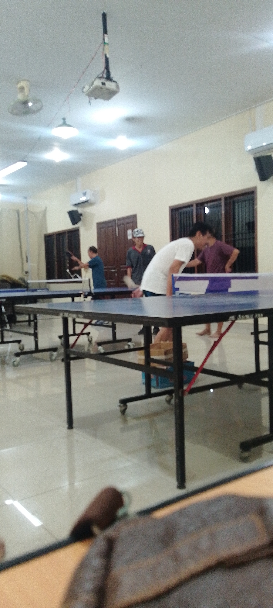 The image depicts an indoor scene, likely a recreational facility or community center, with multiple ping pong tables set up. In the foreground, the edge of a table with a brown object on it is visible, possibly a bag or mat. The environment is well-lit with overhead fluorescent lights. Several individuals are present, engaged in or observing a game of ping pong. One person in a white t-shirt is leaning forward over a table, appearing to play. Another person in a blue t-shirt is visible further back, also near a ping pong table. Two other individuals, one in a dark t-shirt and cap, and another in a burgundy t-shirt, are also present, with the former appearing to be watching. The room has a tiled floor and walls painted in a light color. There are also windows with blinds and wooden doors visible in the background, as well as what appears to be an air conditioning unit mounted on the wall. A projector is suspended from the ceiling. The overall impression is a casual gathering centered around the sport of ping pong. Without further information, the exact location in Kudus, Indonesia cannot be definitively determined from the visual cues alone.