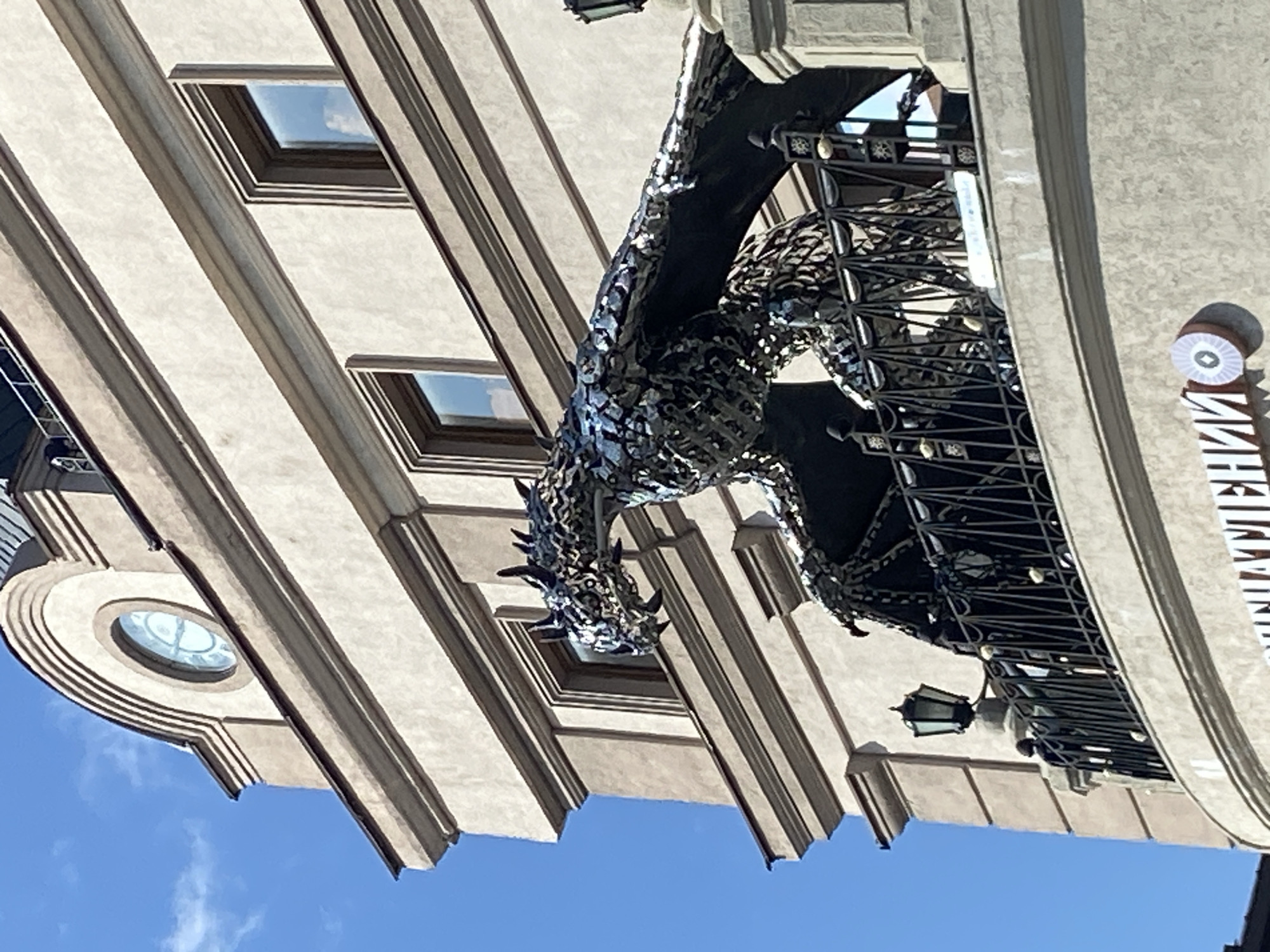 The image captures a close-up, low-angle view of a building facade in Kazan, Russia, featuring a large, metallic dragon sculpture perched on a balcony. The building is light beige with architectural details, including cornices and arched window frames. The dragon sculpture, made of a mosaic of metallic pieces, is detailed with sharp spikes and reptilian features, its wings spread wide. The balcony is adorned with a black metal railing, some sections of which are decorated with star-shaped accents. A black lantern is mounted on a pedestal to the left of the dragon. Below the balcony, a sign with white Cyrillic lettering and a circular emblem is visible on the building's lower level. The sky above is a clear, bright blue with a few wispy white clouds. The scene is lit by natural sunlight, casting shadows and highlighting the textures of the building and the sculpture. The overall impression is one of an artistic and somewhat imposing presence within an urban setting.