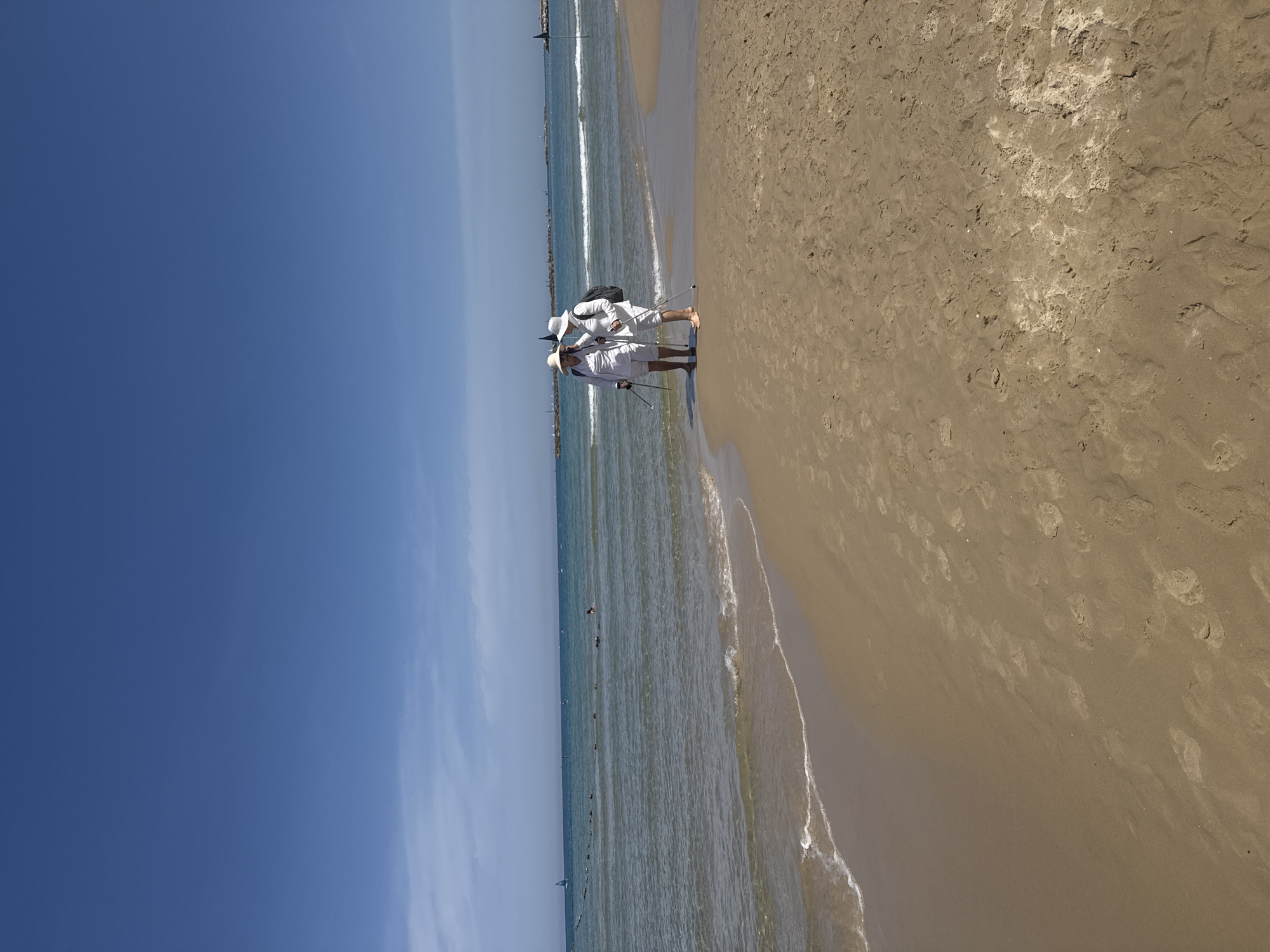 Two people in white clothing, including hats, walk barefoot on the beach, each using a walking stick. The sky is clear blue. The beach is sandy with footprints. The water is calm with small waves. A safety barrier is visible in the distance. The location is Tel-Aviv beach, Israel.